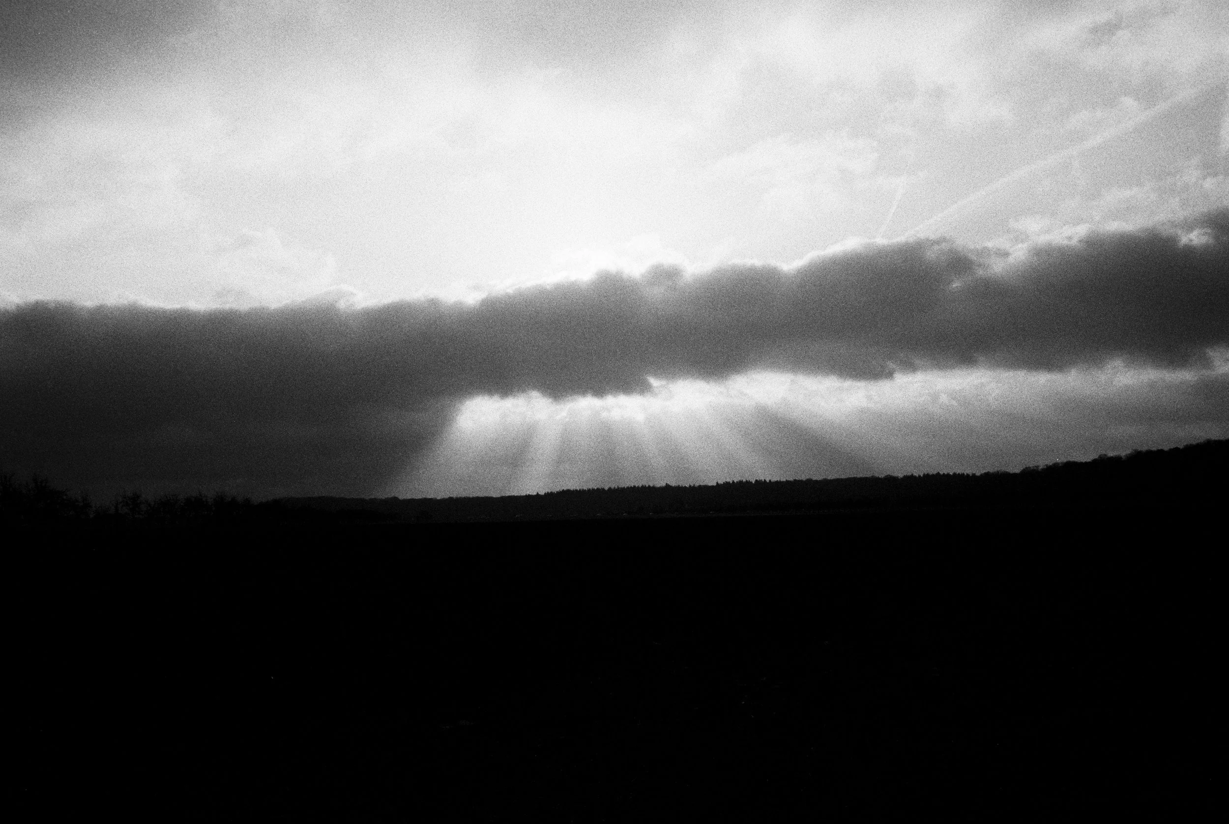 Schwarzer Himmel mit Wolken und Sonnenstrahlen, die durch die Wolken scheinen, über einer Landschaft im Hintergrund.