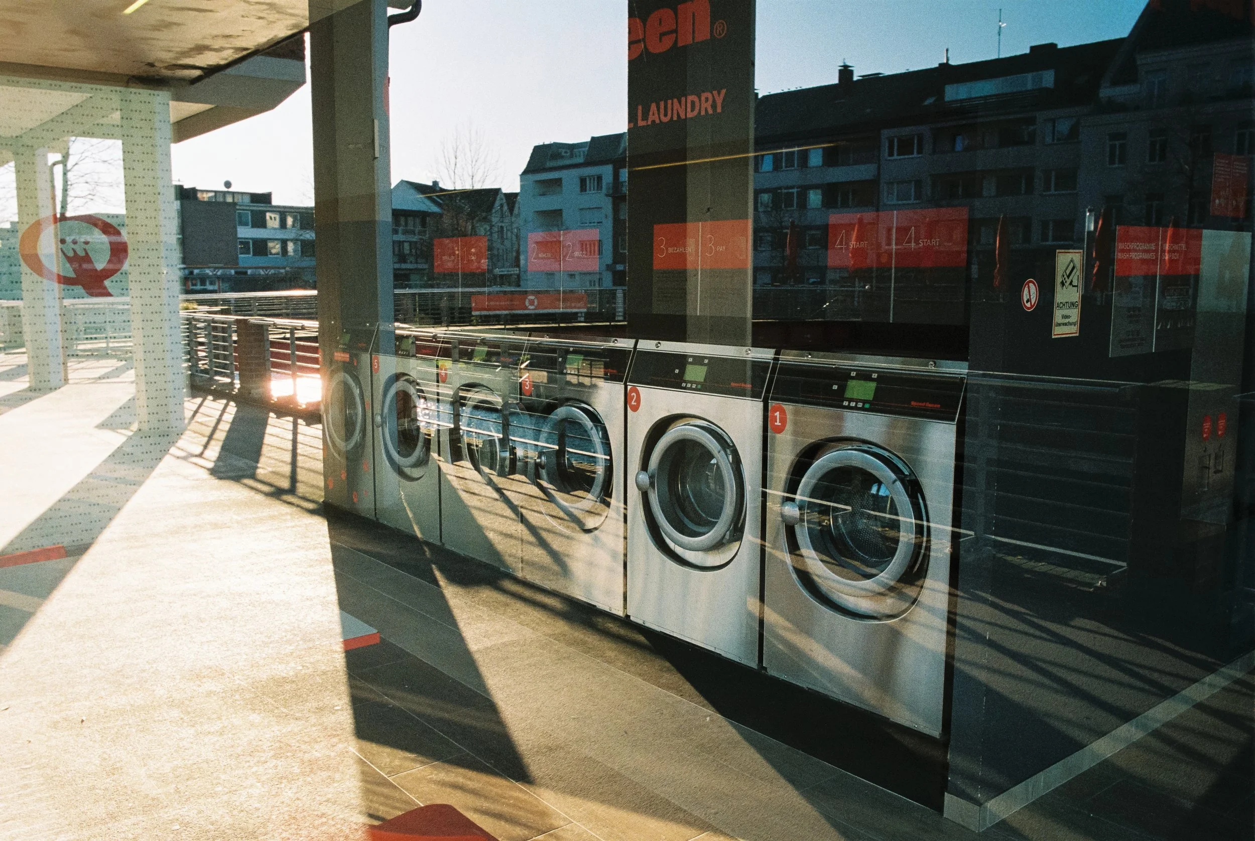 Waschmaschinen in einer Wäscherei, sichtbar durch die Fenster. Gebäude und ein klarer Himmel im Hintergrund, reflektiert im Fenster.
