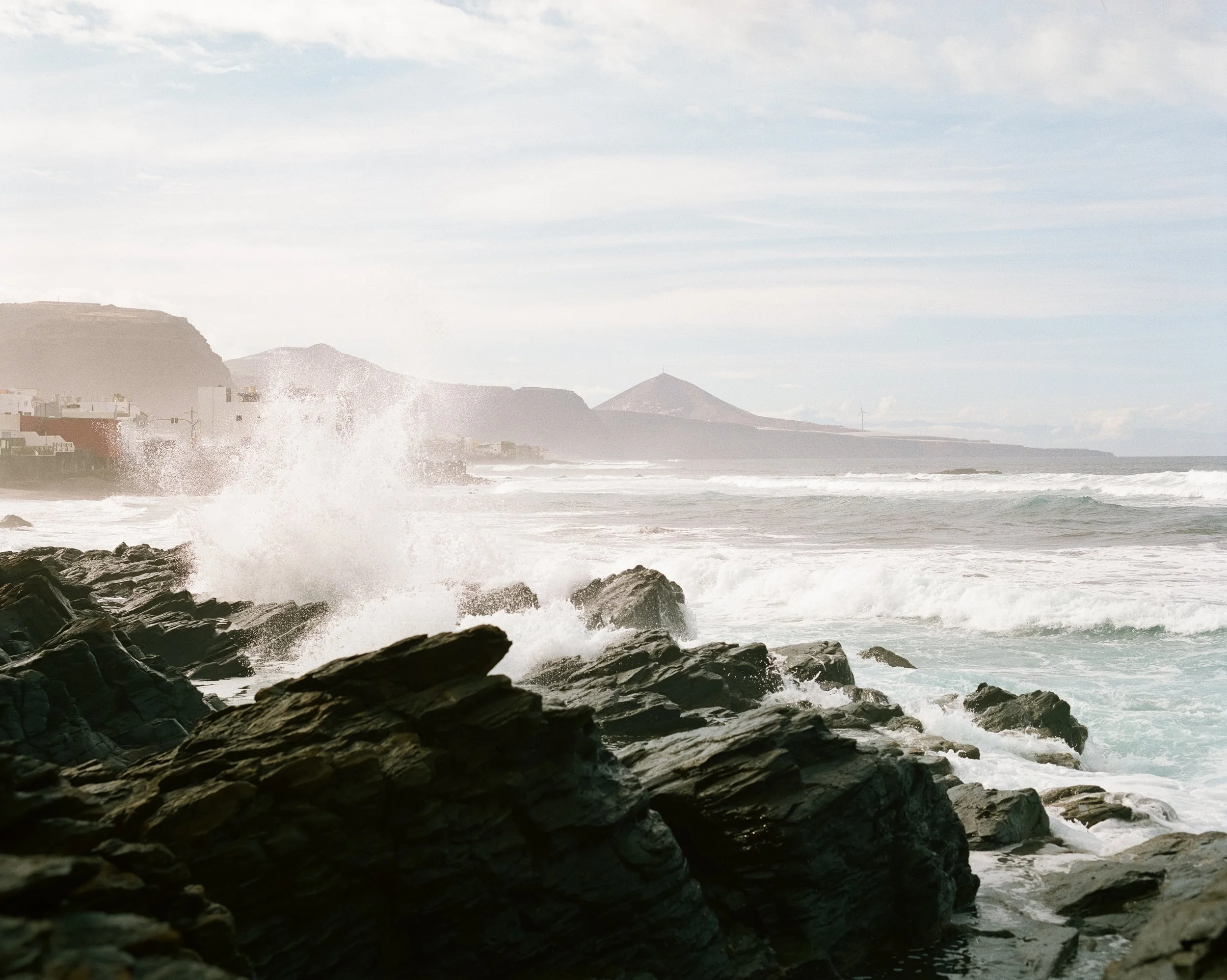 Auf den Felsen vor der Küste schlagen die Wellen. Es ist ein stürmischer Tag am Meer mit Wolken am Himmel und einem entfernten Landmassiv im Hintergrund.