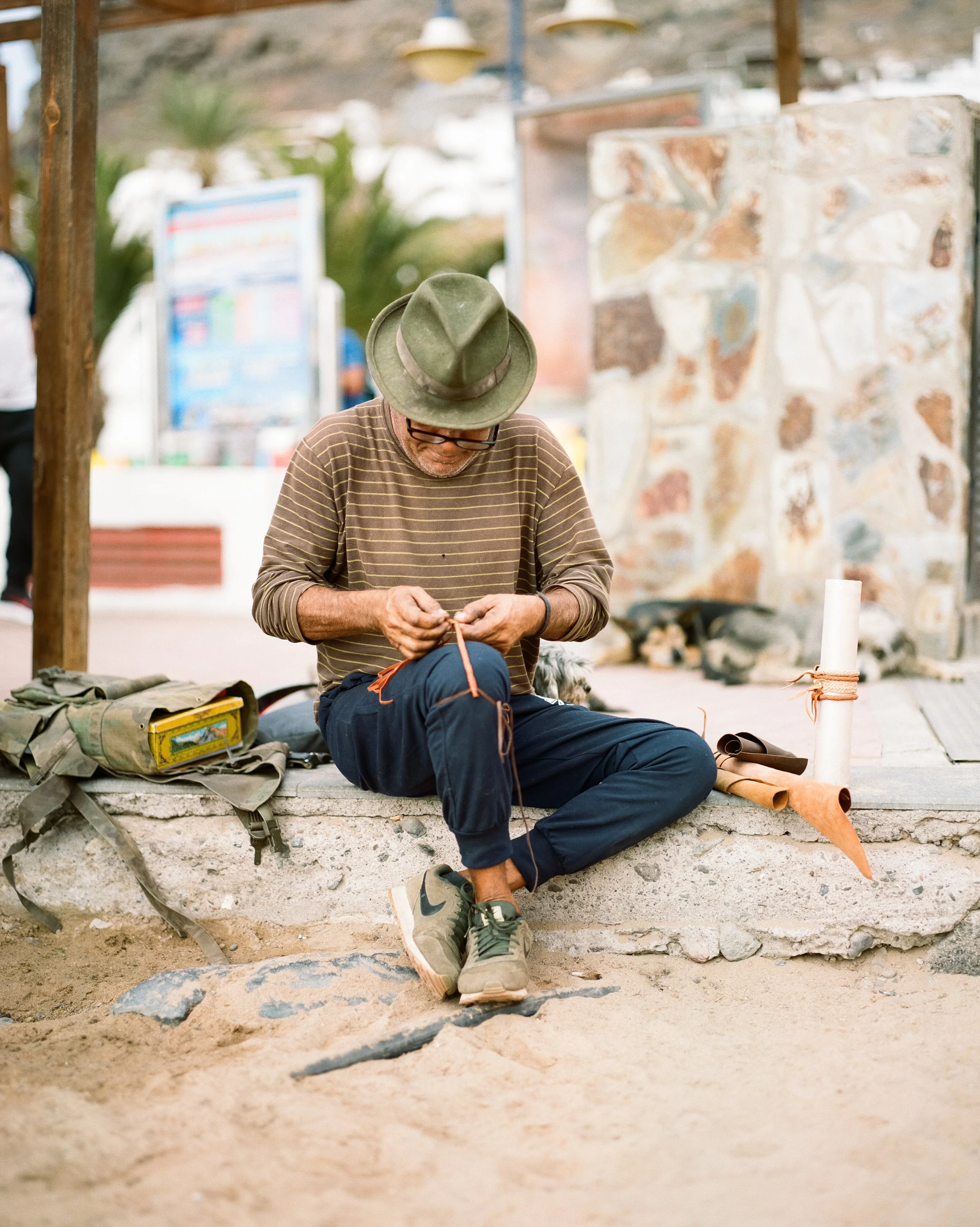 Ein Mann sitzt auf einer Steinbank, trägt einen grünen Hut, eine braune gestreifte Hemd, dunkle Hosen und Sneakers und arbeitet an einem Handwerksprojekt mit einem roten Faden. Er sitzt in einem öffentlichen Raum mit Sandboden, einer Mauer im Hinterg