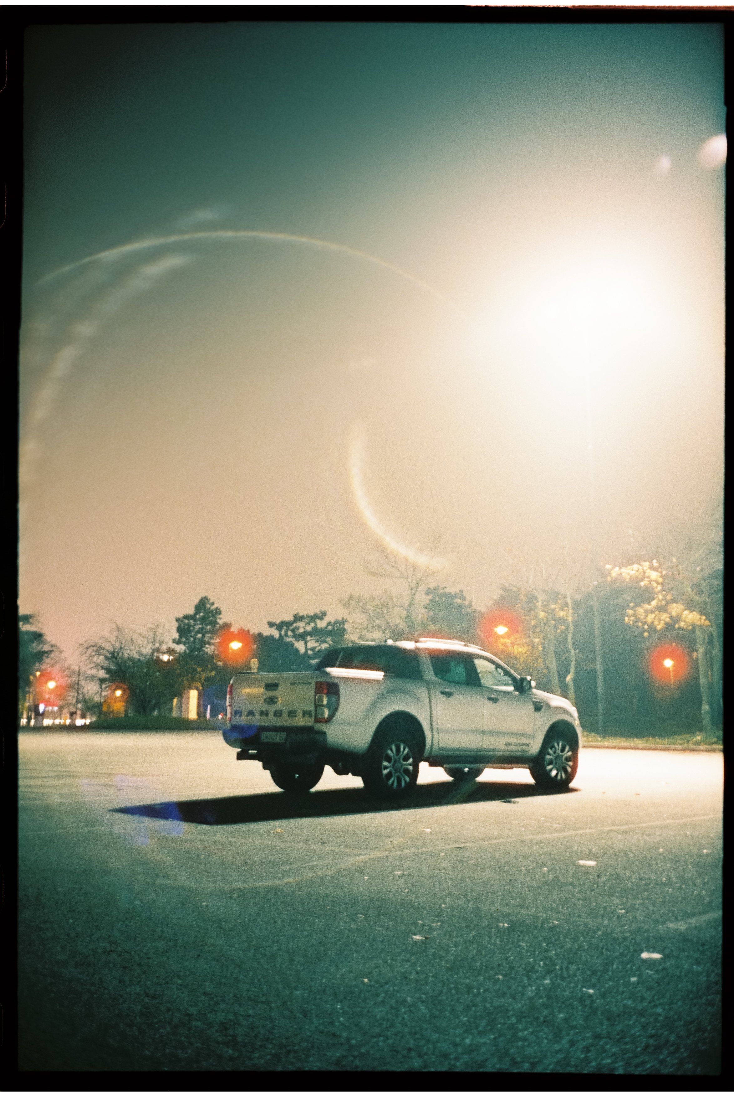 Ein weißer Pickup-Truck steht auf einem leeren Parkplatz bei Nacht mit streetlights im Hintergrund. Der Himmel ist neblig und es gibt Lichtspuren am Himmel, die auf Langzeitbelichtung hinweisen.