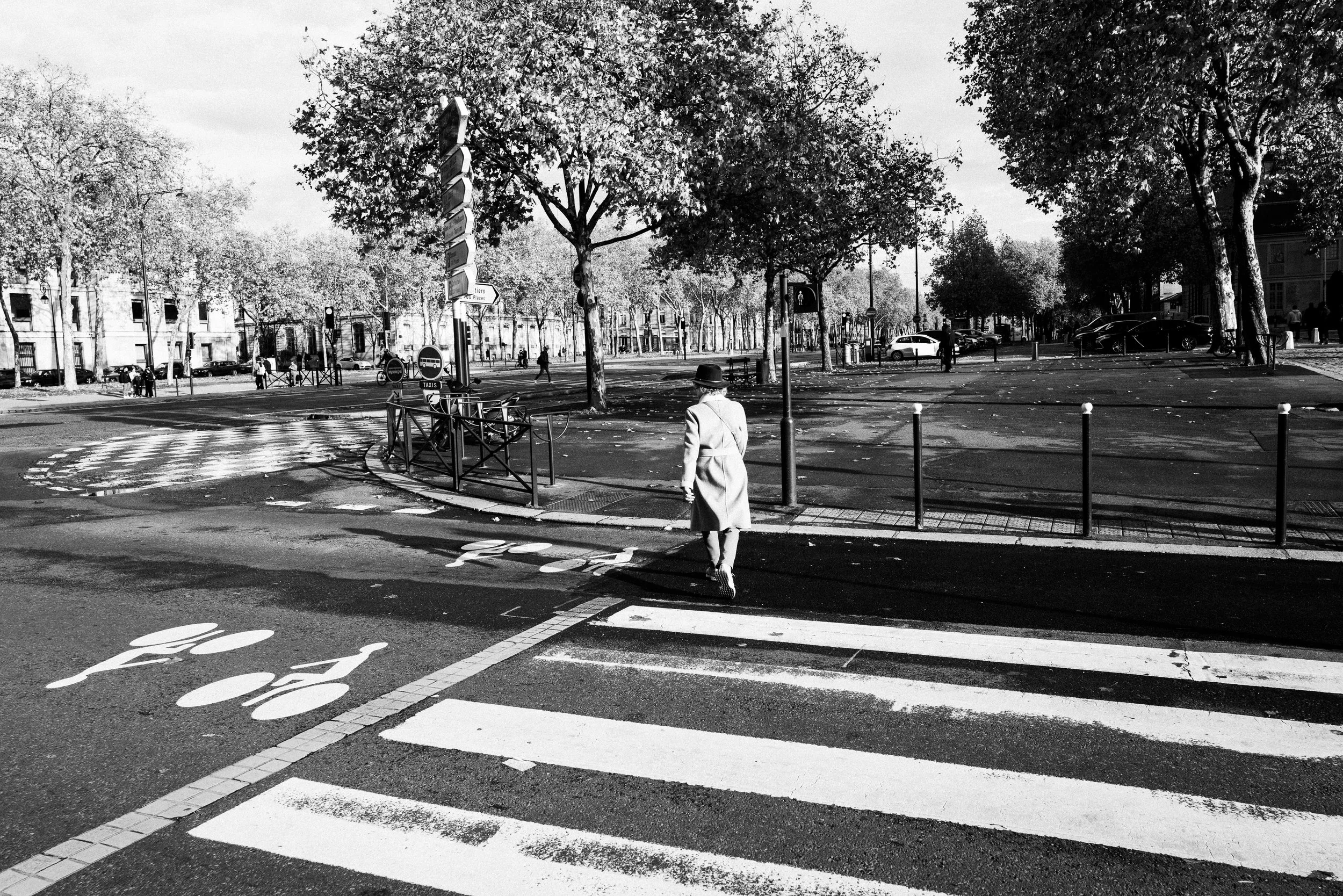 Ein Mann in einem Mantel und Hut geht über einen Zebrastreifen auf einer Stadtstraße, im Hintergrund Bäume und Gebäude, alle im Schwarz-Weiß-Filter.