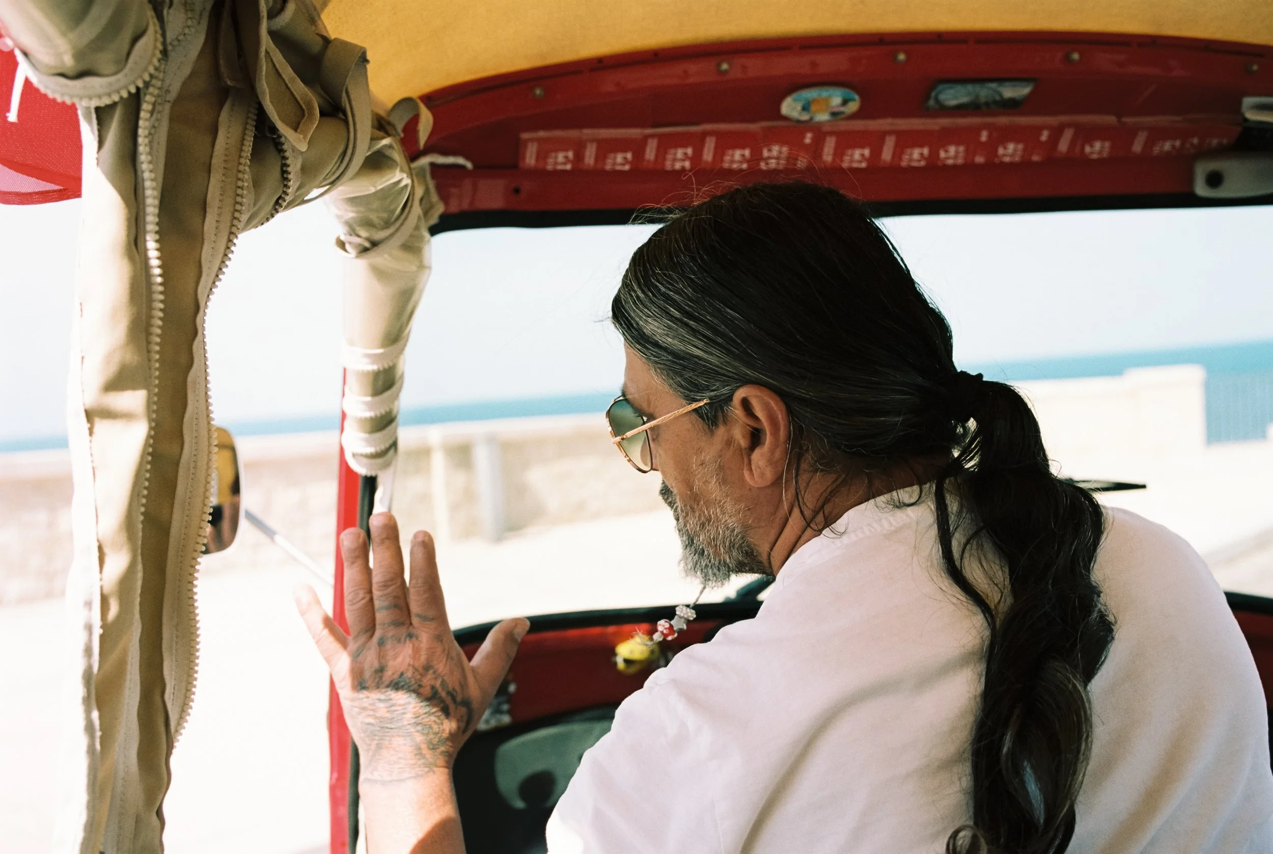 Ein Mann mit langen, dunklen Haaren, Brille und Tattoos sitzt in einem roten Fahrzeug und winkt. Im Hintergrund ist der Himmel und das Meer sichtbar.