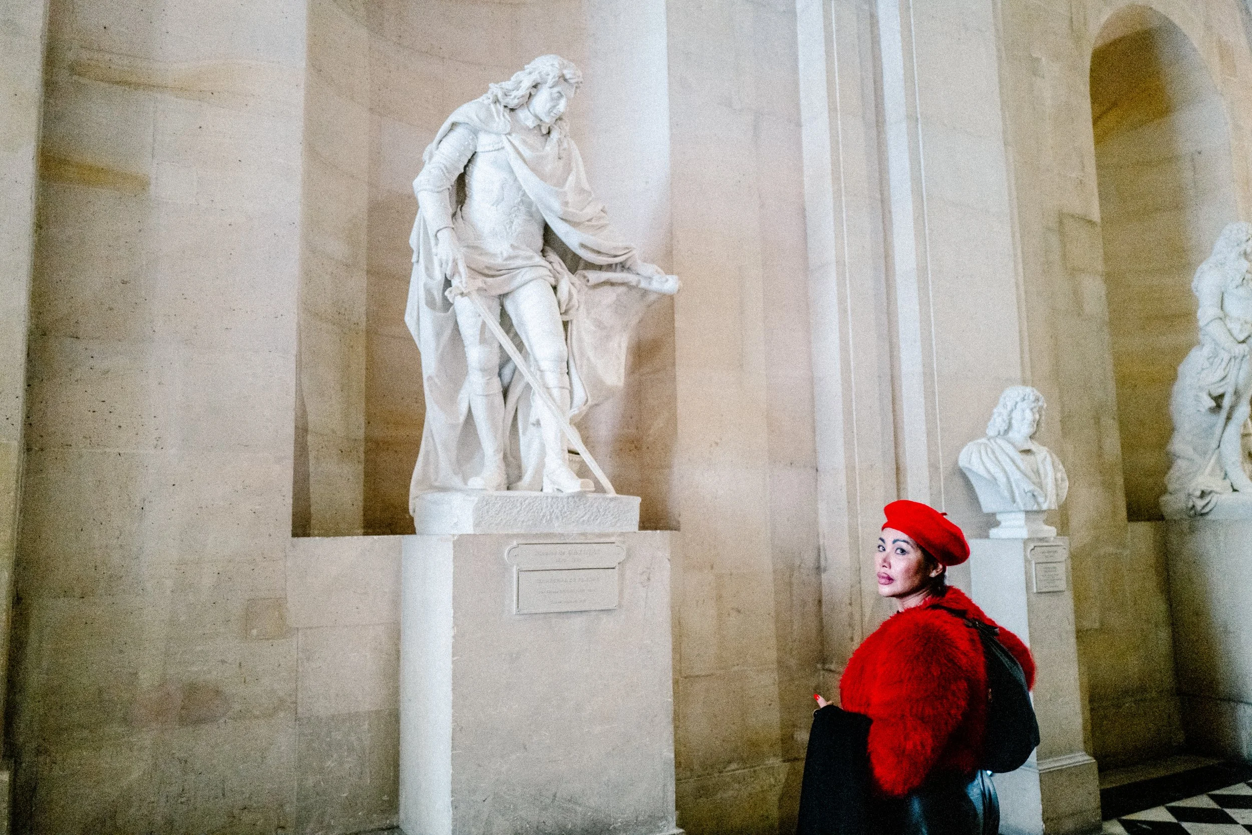 Eine Frau in rotem Hut und roter Pelz über einem schwarzen Mantel blickt zu einer Skulptur in einem Museum.