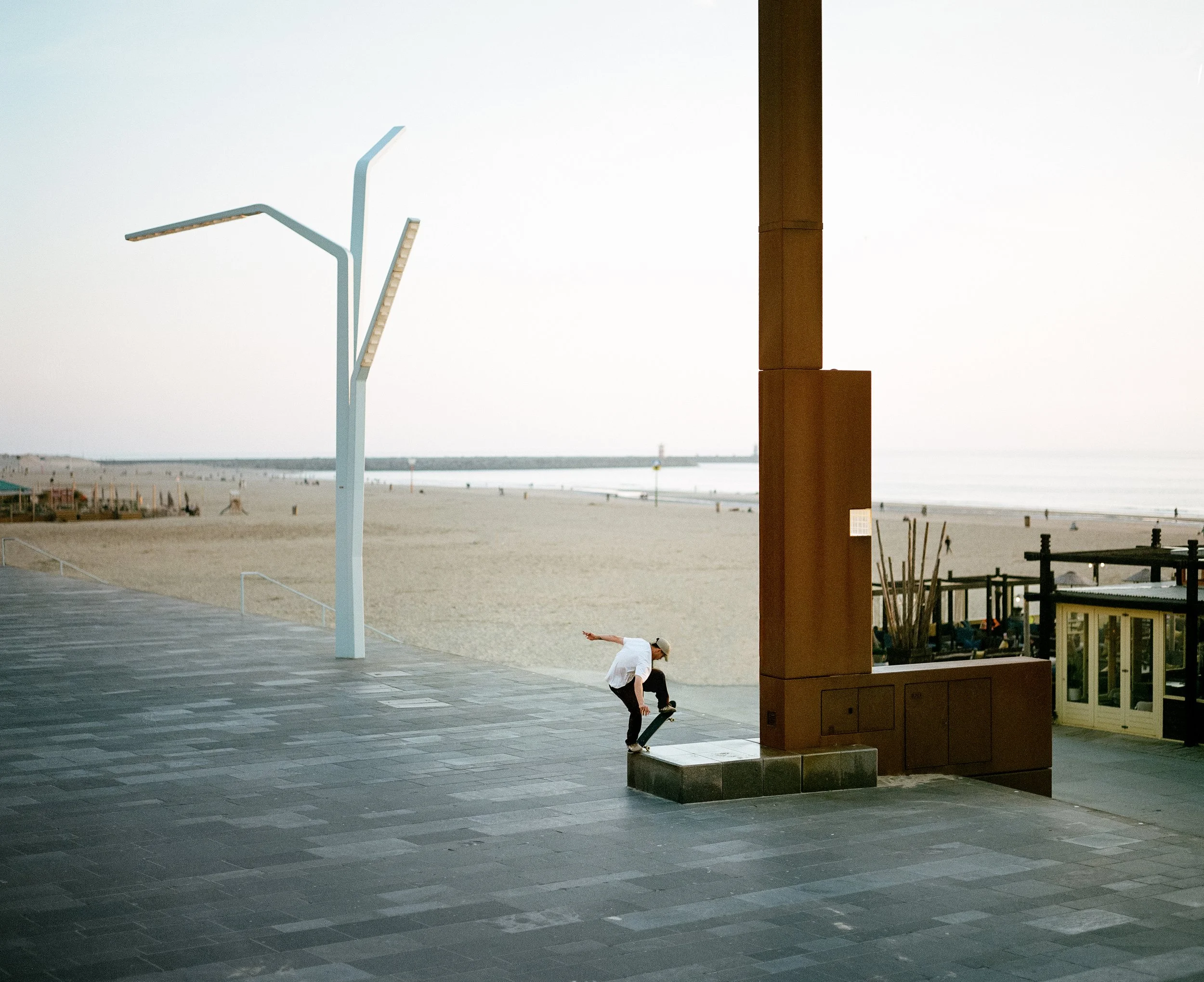 Ein Skateboarder macht einen Trick auf einem Bordstein an einem Strand bei Sonnenaufgang, im Hintergrund leere Strandfläche und Meer.
