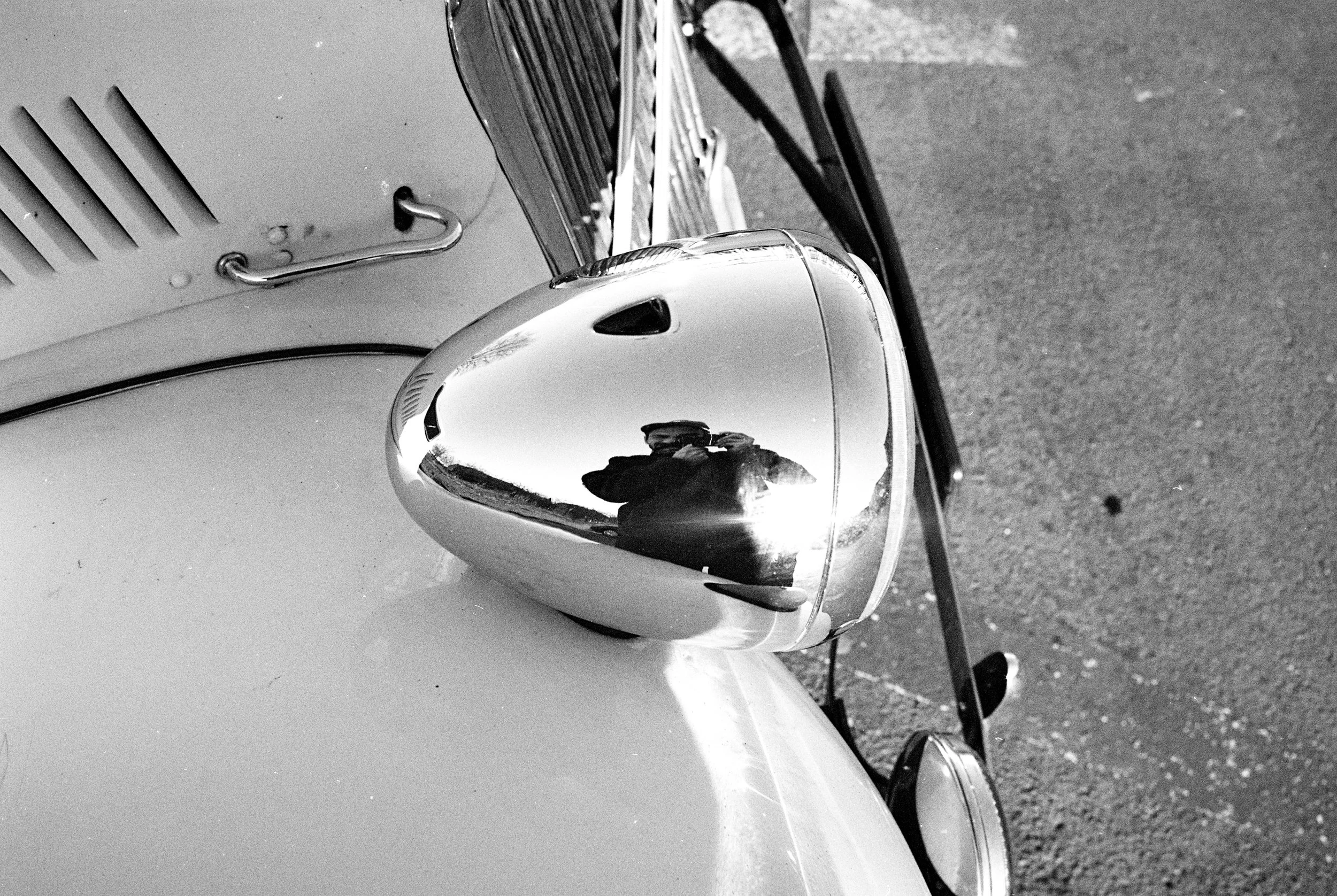 Nahaufnahme des vorderen Teils eines alten Autos, einschließlich Scheinwerfer und Spiegel, in Schwarz-Weiß fotografiert, mit einer reflektierenden Oberfläche im Spiegel, die eine Person zeigt, die den Fotonah gemacht hat.