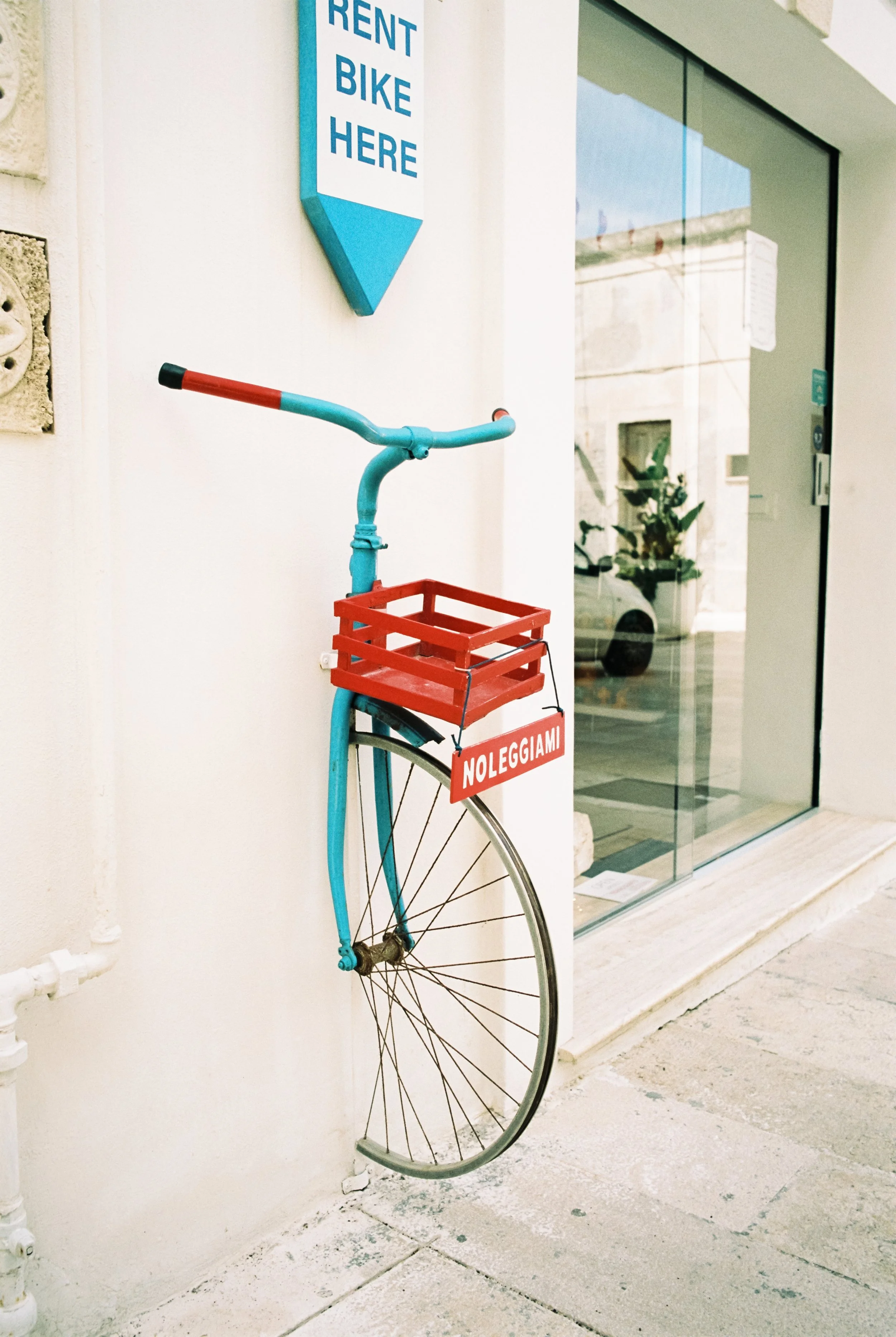 Ein bemalter Fahrradkorb hängt an einer weißen Wand. Es gibt ein Schild darüber, das sagt 'RENT BIKE HERE'. Das Fahrrad ist rot und blau, mit einem roten Korb und einem Schild, das 'NOLEGGIAMI' sagt, was auf Italienisch 'Miete mich' bedeutet.