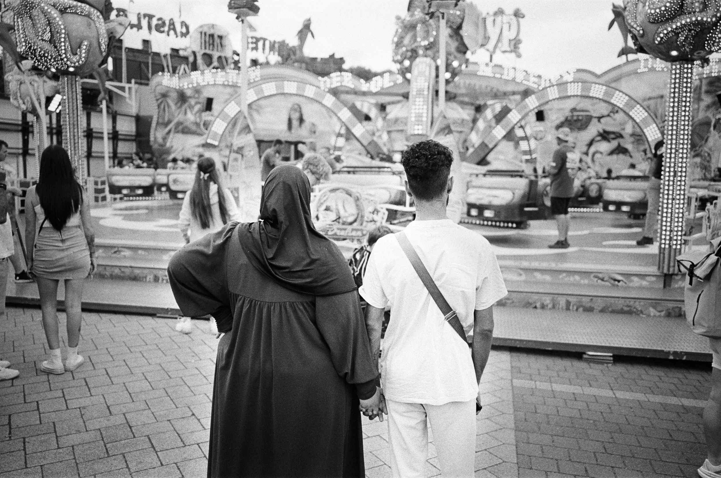 zwei Menschen, die auf einem Jahrmarkt stehen, halten sich an den Händen, im Hintergrund Karussell mit fahrenden Personen, andere Besucher