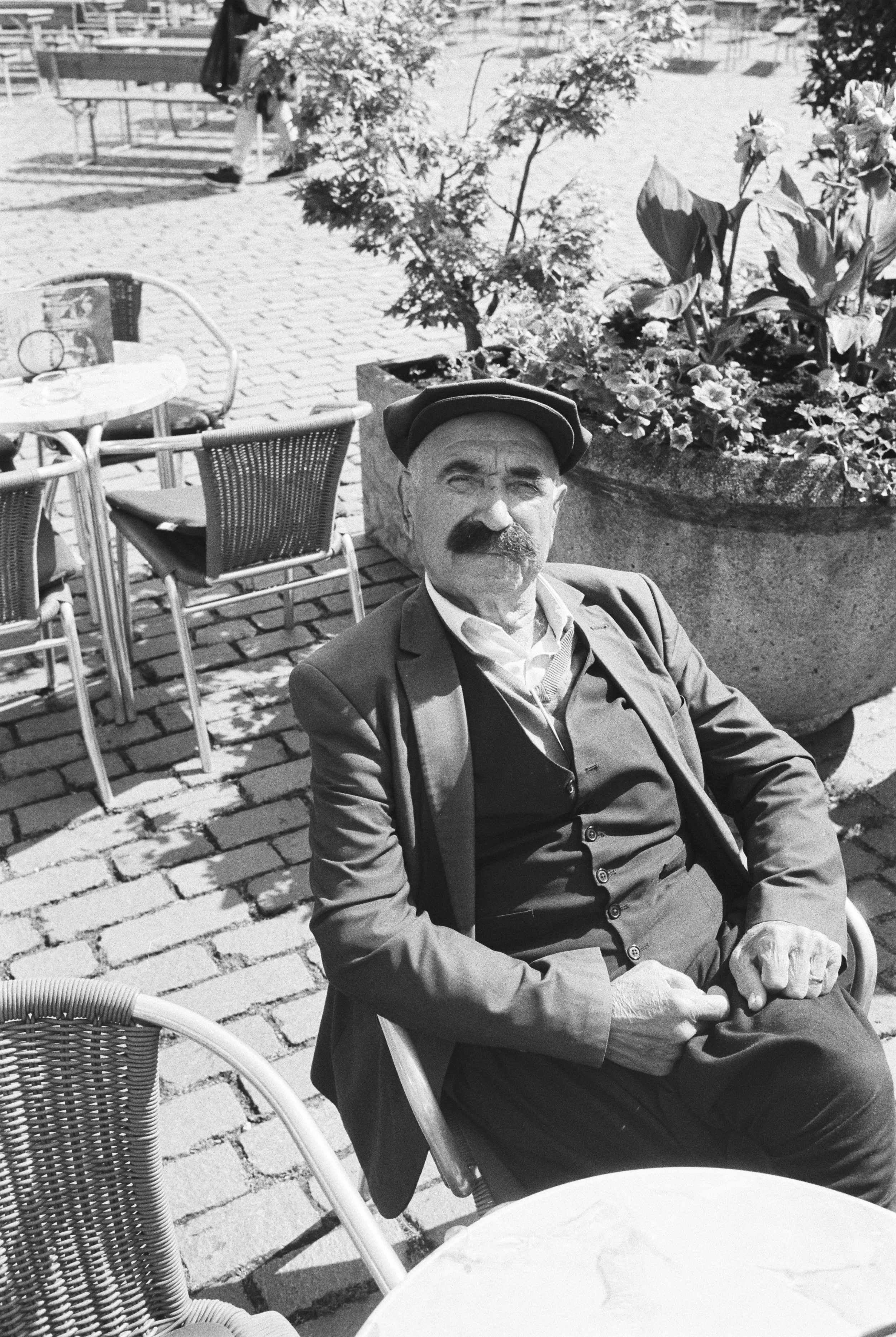 Ein Mann mit Schnurrbart und Hut sitzt auf einer Terrasse im Freien, umgeben von Tischen und Stühlen, in einer Stadt mitpflanzen auf einem Pflanzkübel im Hintergrund.