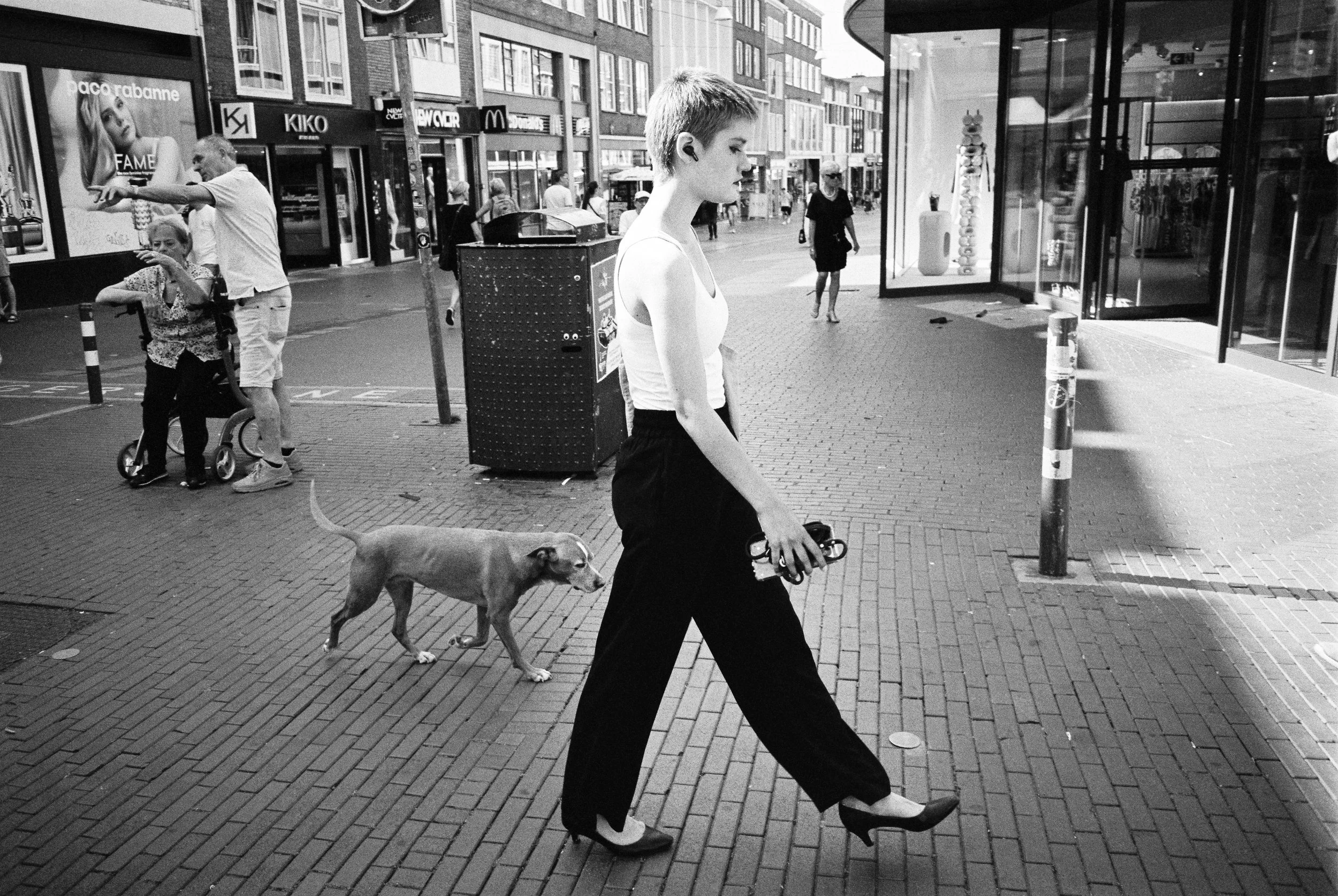 Eine junge Frau mit kurzem Haar, weißen Top und schwarzen Hosen geht auf einer Stadtstraße mit einem Hund. Im Hintergrund sind Einkaufsgeschäfte und Passanten.