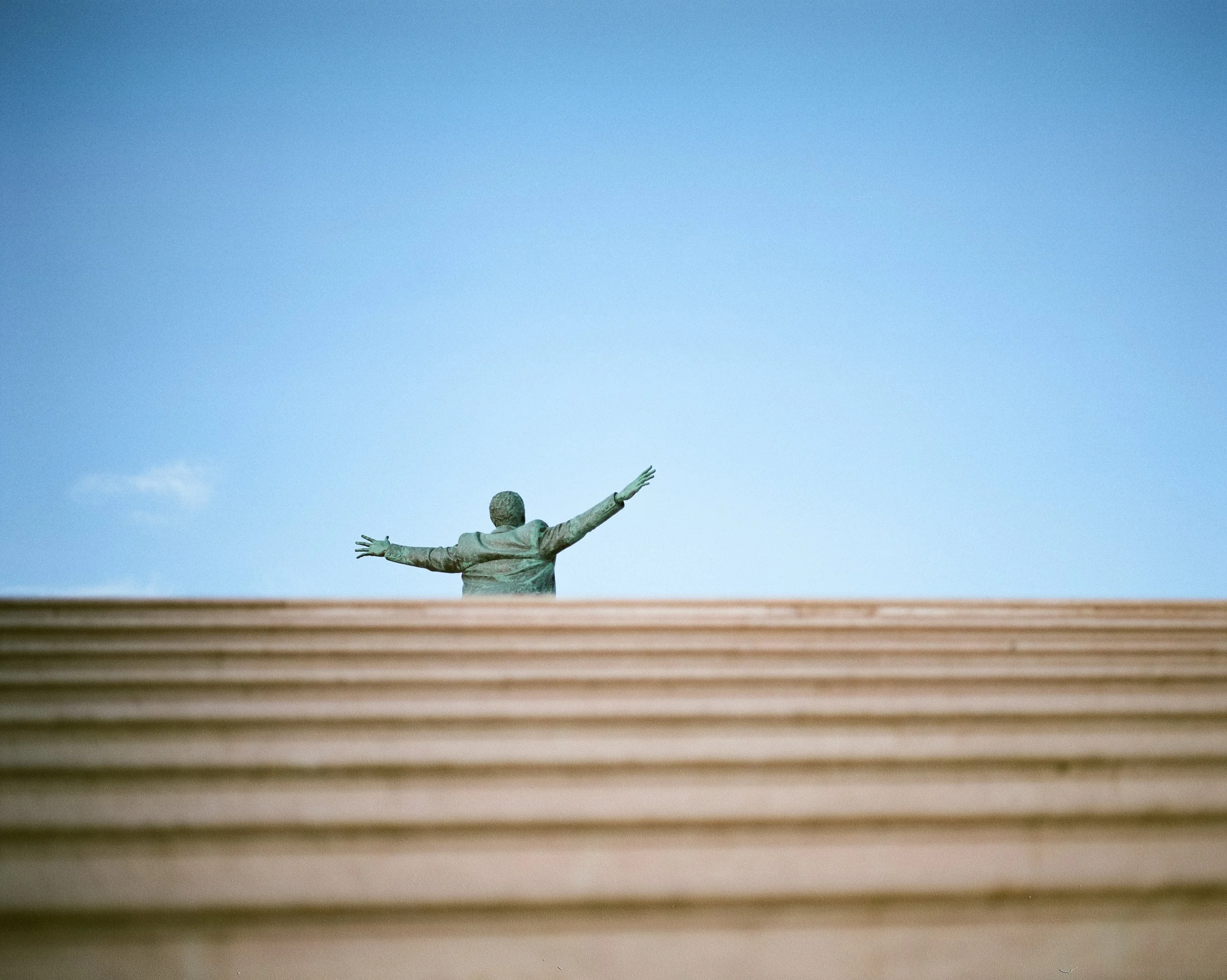 Bronzefigur eines Menschen mit ausgebreiteten Armen auf einem Podest, Blick nach oben, gegen blauen Himmel