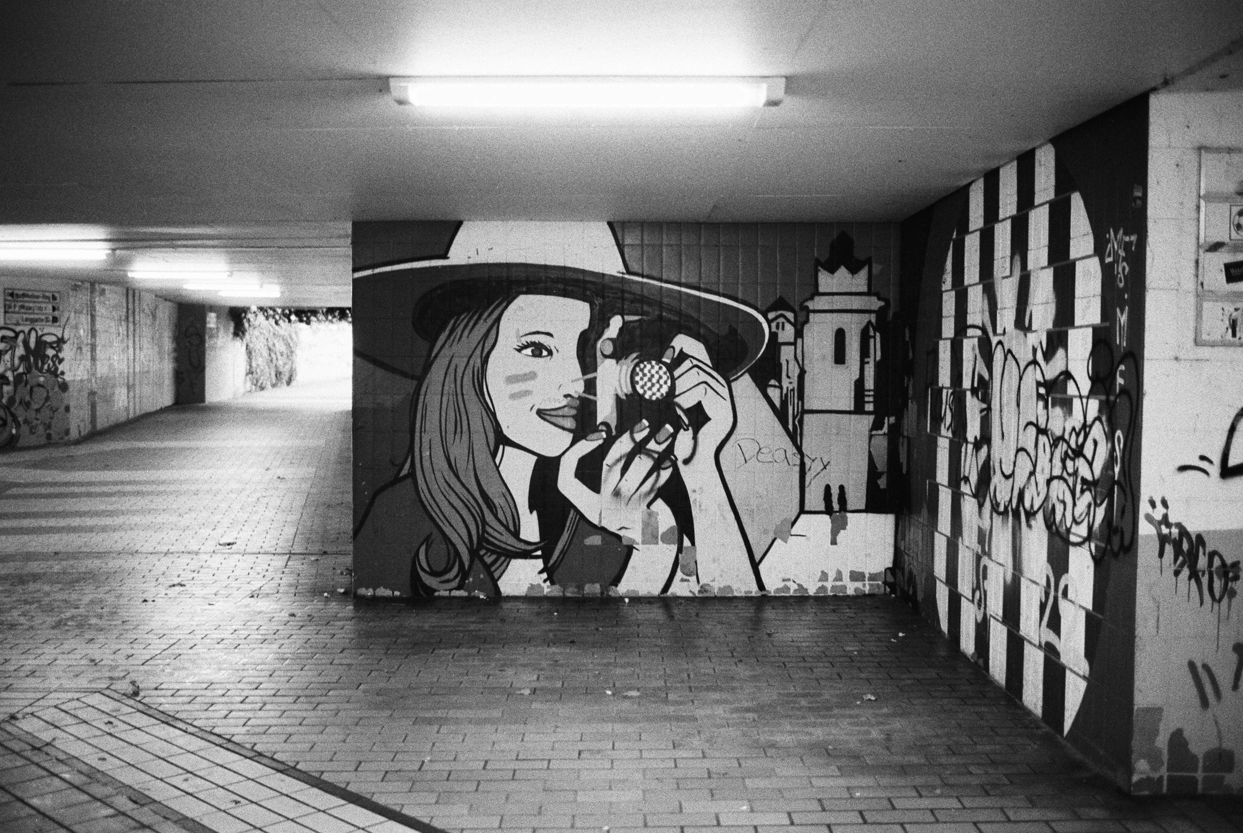 Graffiti einer Frau mit Hut, die eine Kamera hält, in einem unterirdischen Tunnel mit Graffiti an den Wänden.