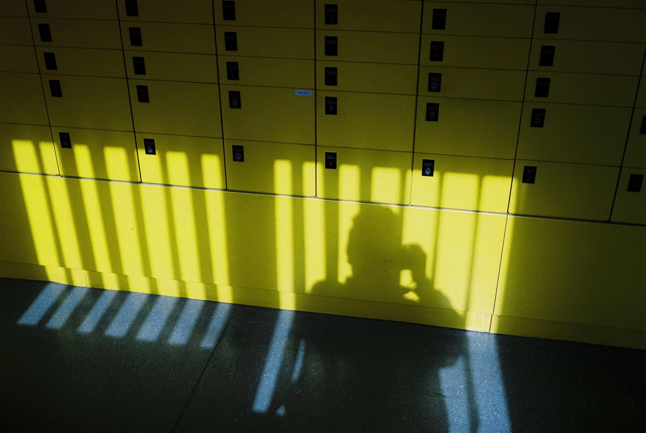 Schatten eines Menschen, der mit einem Mobiltelefon fotografiert, wird auf eine gelbe Wand mit Postfächern projiziert, mit Sonnenstrahlen, die durch einen Gittervorhang fallen.