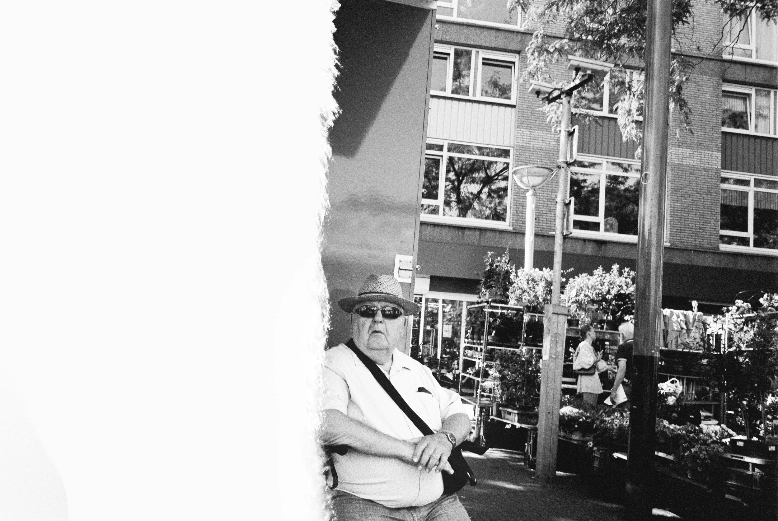 Ein älterer Mann trägt einen Strohhut, Sonnenbrille, weißes Hemd und eine schwarze Tragetasche auf der Schulter, steht vor einem Blumenstand in einer städtischen Umgebung mit Gebäuden und anderen Personen im Hintergrund.