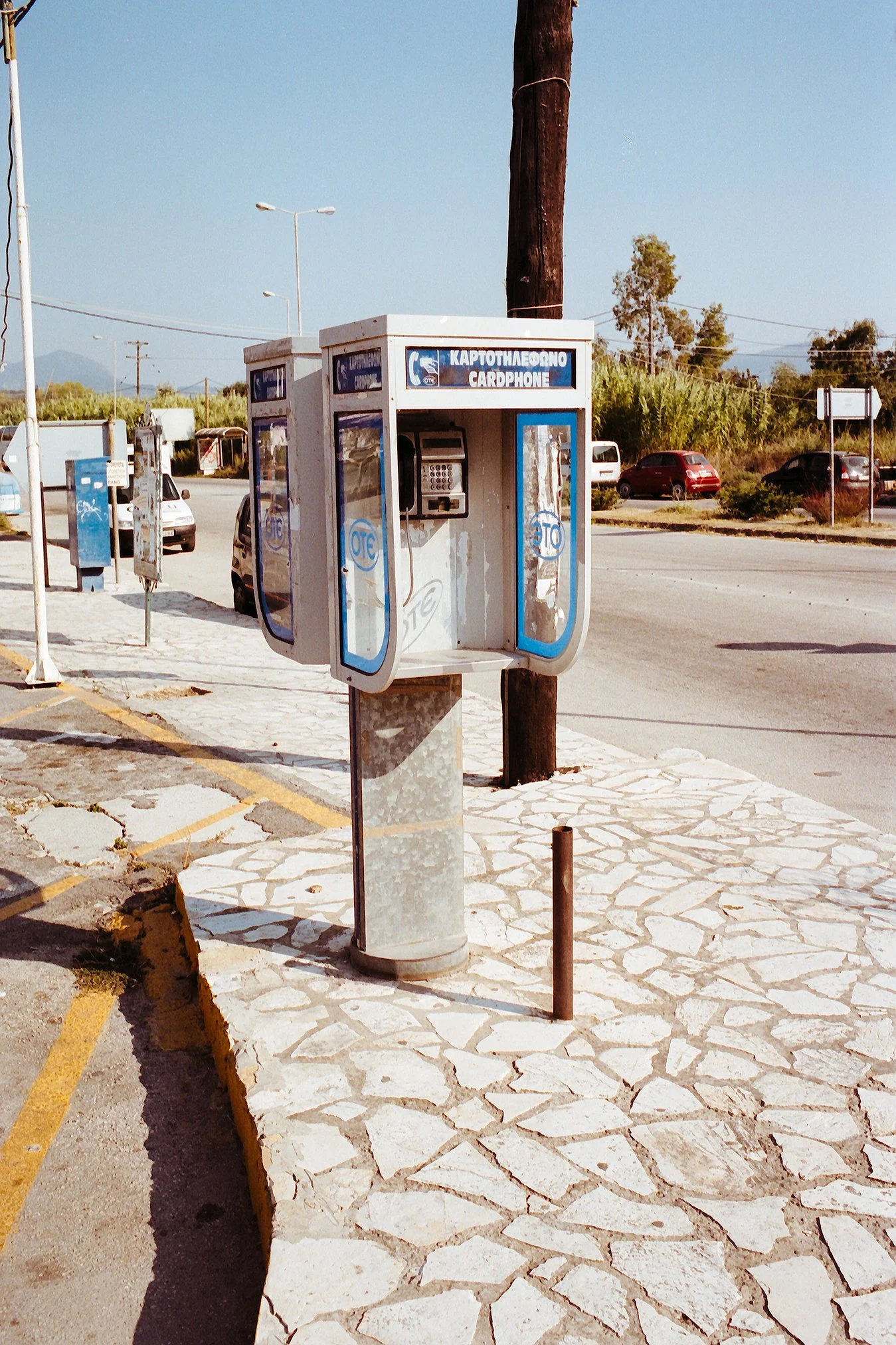 Ein Alphanumerischer Kartenautomat (Telefonautomat) an einer Straßenecke auf einem gepflasterten Bürgersteig, im Hintergrund sind parkende Autos und Bäume zu sehen.