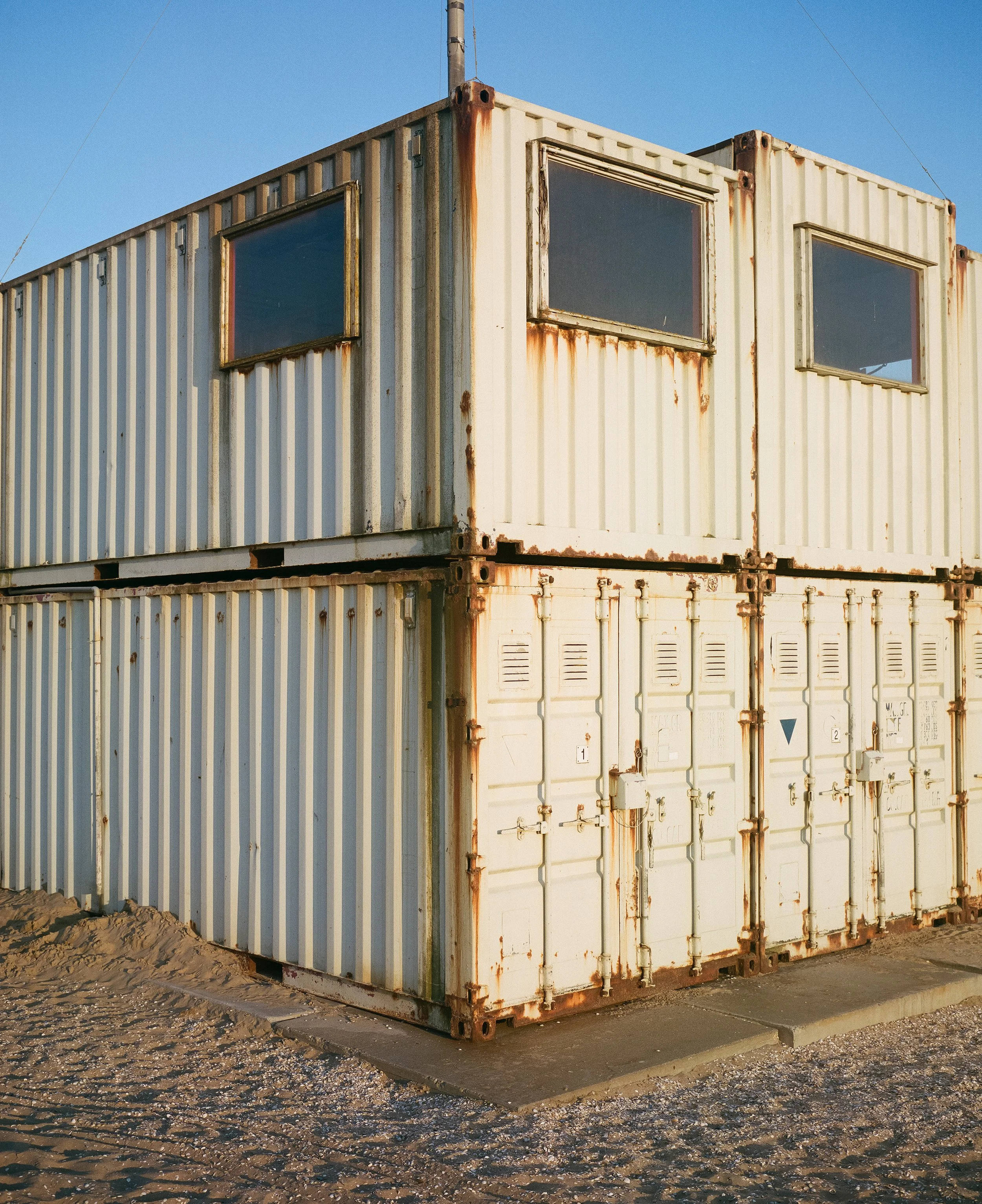 Zwei gestapelte alte, verrostete, weiße Container auf Dreckboden bei Sonnenuntergang mit klarem blauen Himmel.