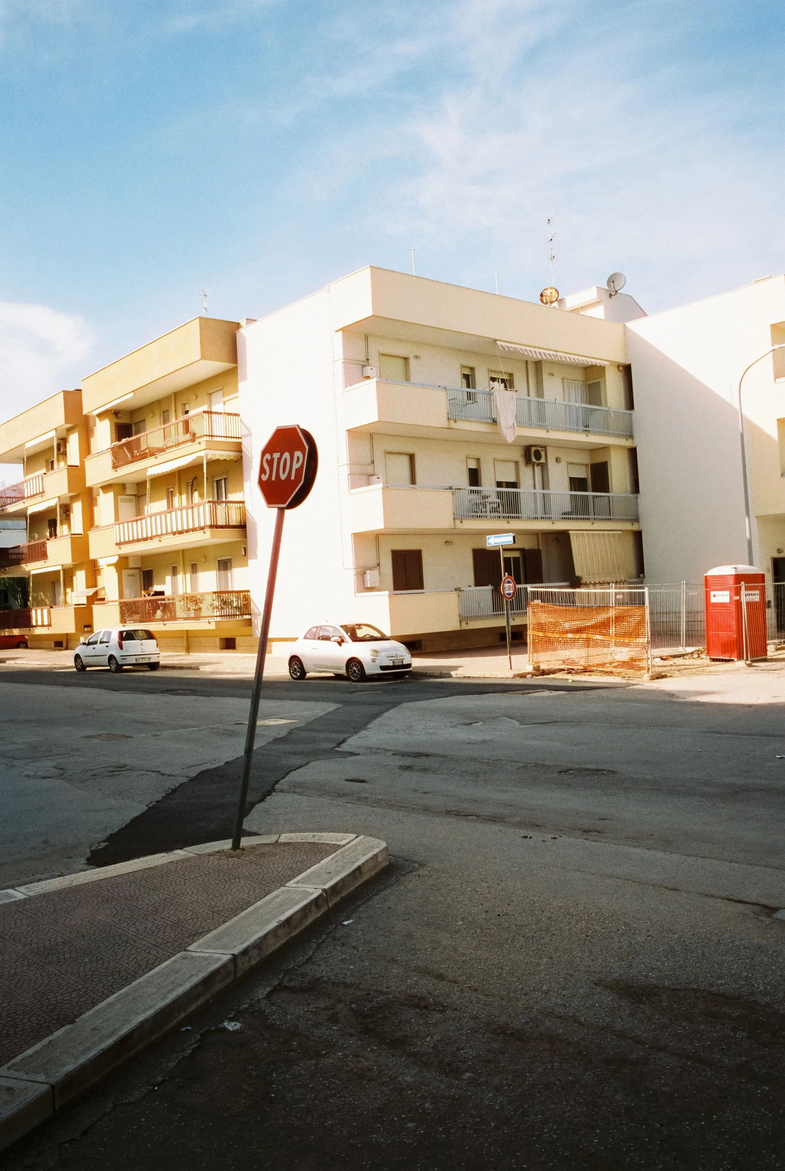 Straße mit einem umgeknickten Stoppschild, mehrstöckige Gebäude mit Balkonen, geparkte Autos, Bauzaun und eine rote Telefonzelle