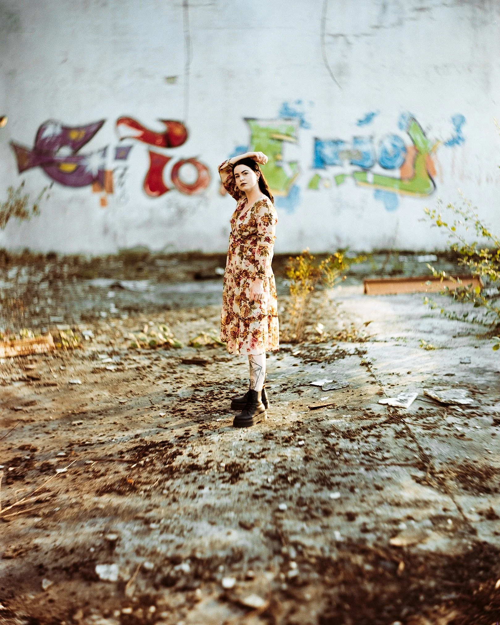 Eine Frau in einem floralen Kleid und schwarzen Stiefeln steht vor einer mit Graffiti bemalten Wand, umgeben von Unkraut und Schmutz auf dem Boden.