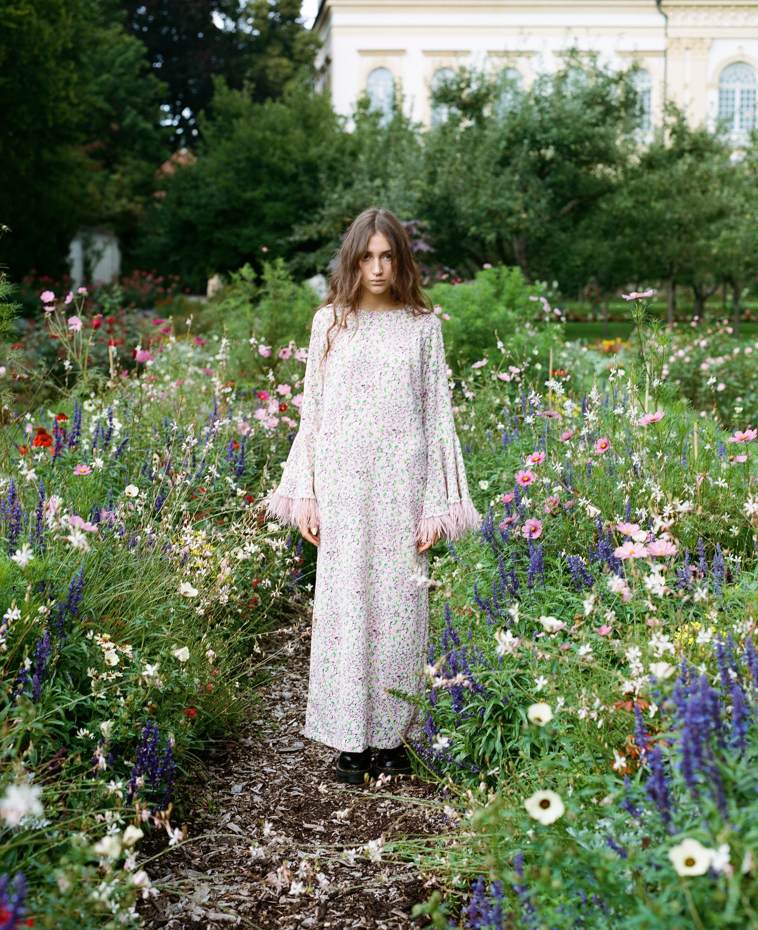 Junge Frau steht auf einem Pfad in einem Blumenfeld, umgeben von bunten Blumen, mit einer historischen Gebäudefront im Hintergrund.