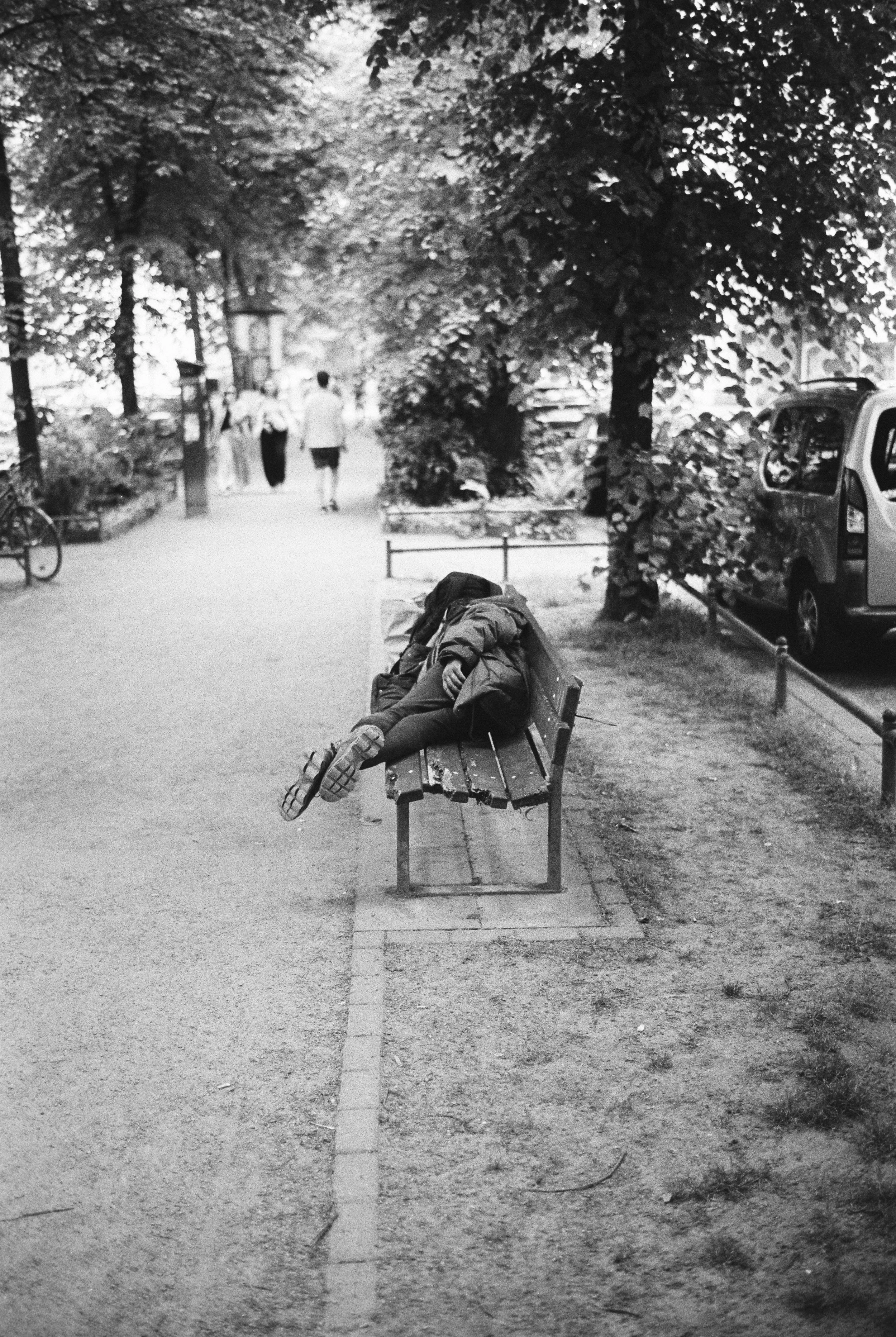Ein schlafender Kind auf einer Parkbank in einem Baumpark, umgeben von Bäumen, mit Menschen im Hintergrund und einem geparkten Auto rechts.