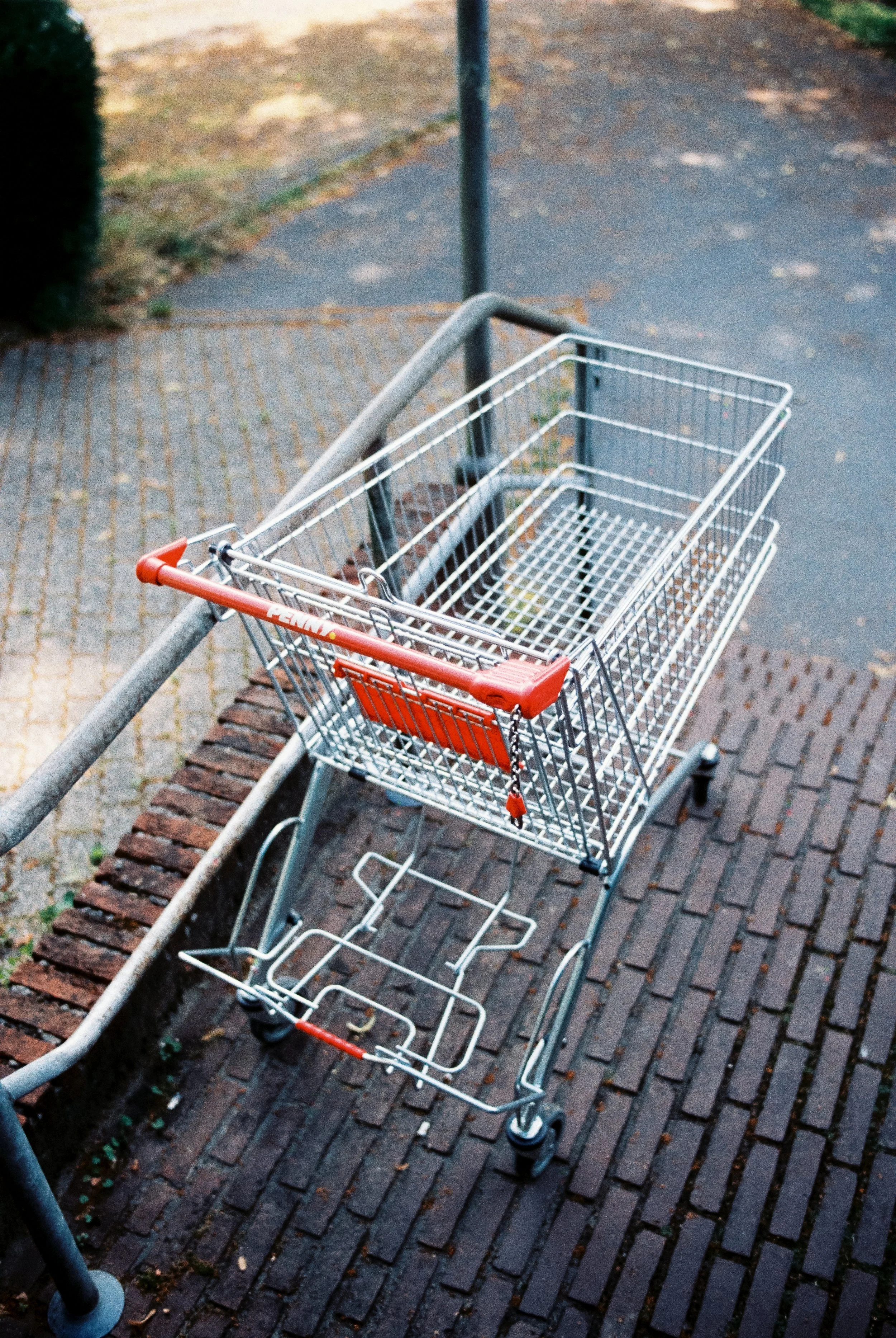 Leeres Einkaufswagen auf gepflastertem Boden neben einem Gehweg mit Asphaltstücken und einem grünen Busch im Hintergrund.
