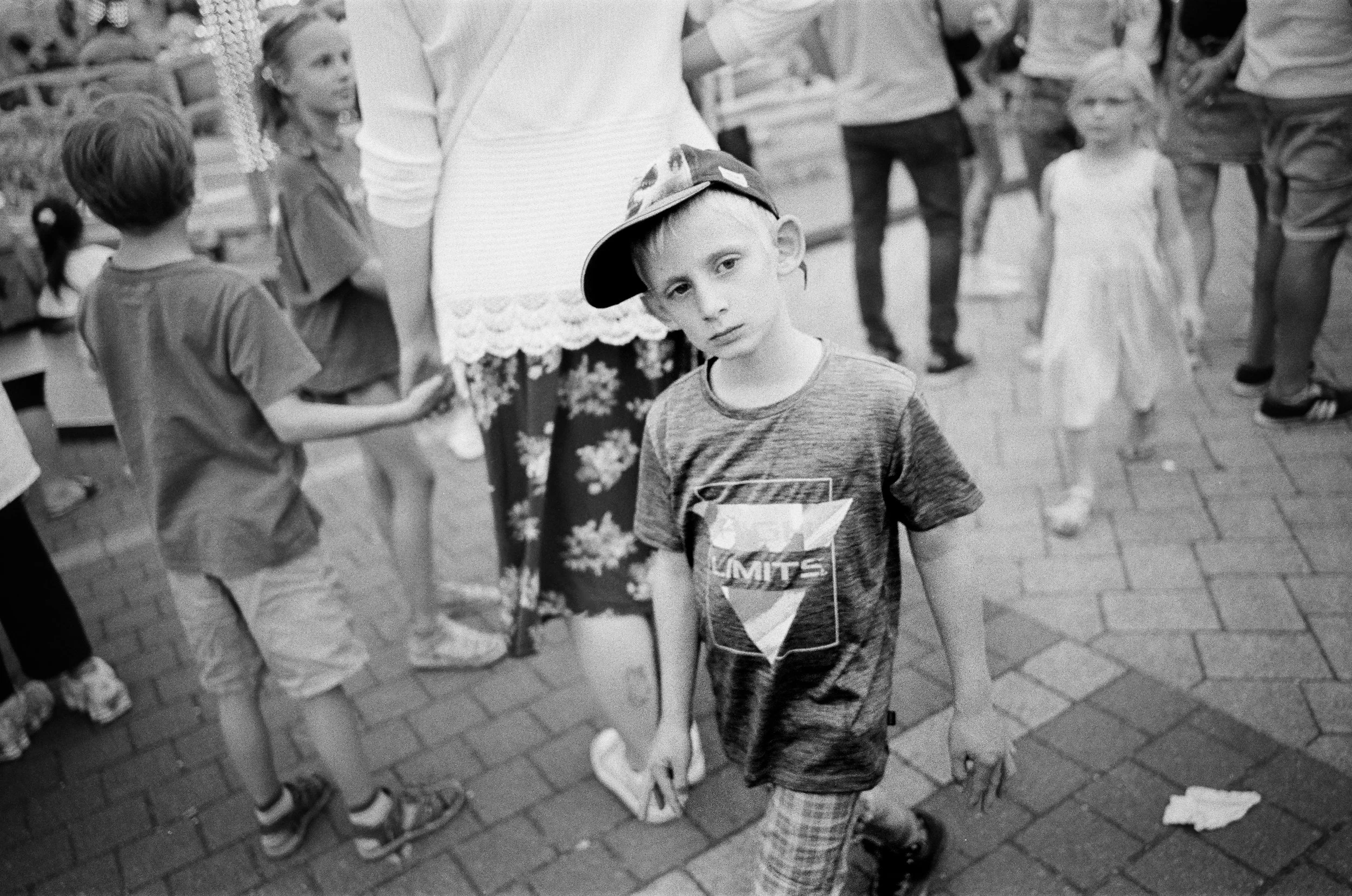 Ein Junge trägt ein T-Shirt mit einem Grafikdesign und eine Mütze schräg auf dem Kopf, steht inmitten einer Menschenmenge auf einem Gehweg.