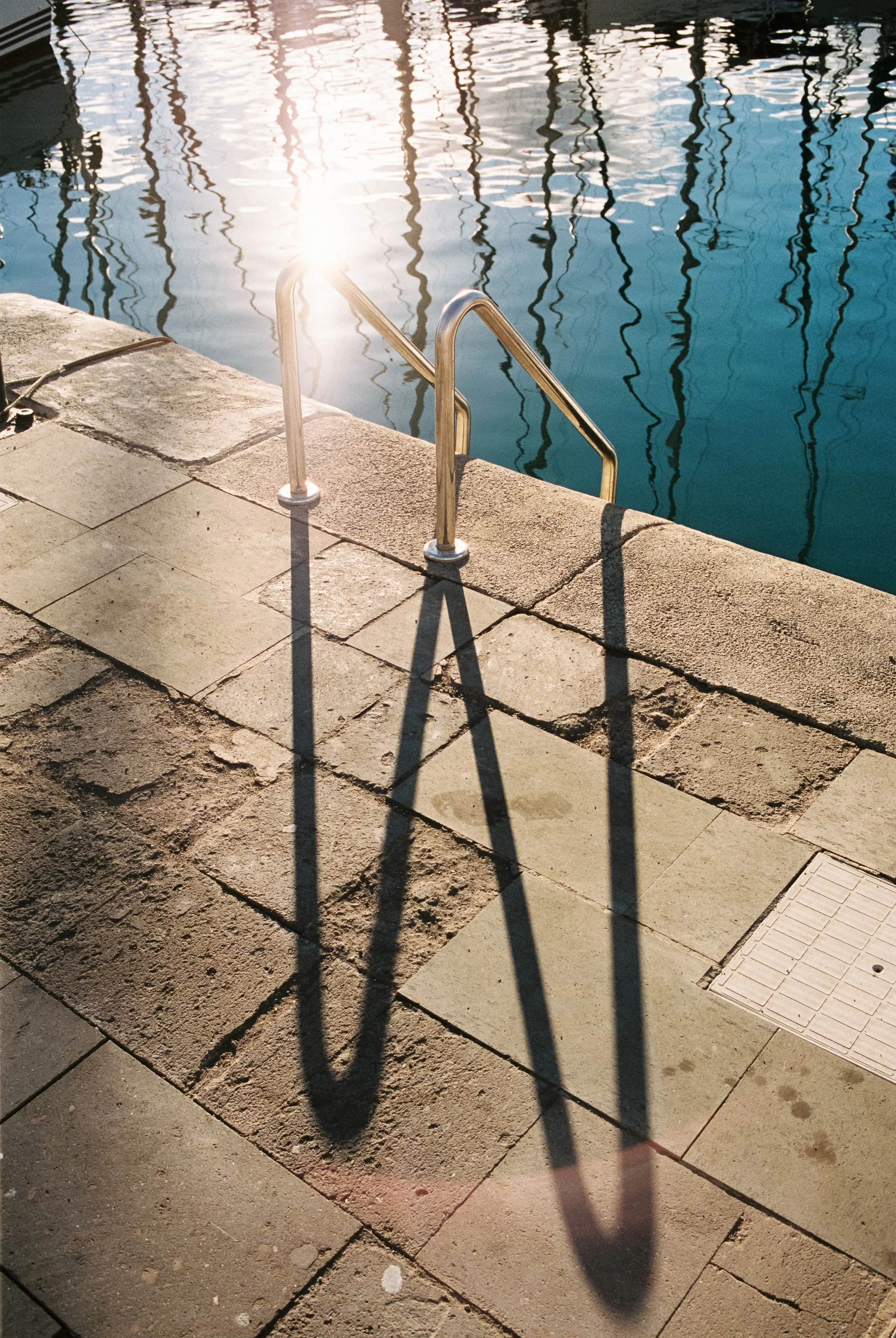 Blick auf das Becken eines Bootshafens mit einem Treppenabsatz, der von der Sonne beleuchtet wird. Die Leiter aus Metall führt ins Wasser, während die Schatten der Leiter auf den Boden fallen. Im Hintergrund spiegeln sich Masten von Booten im Wasser.