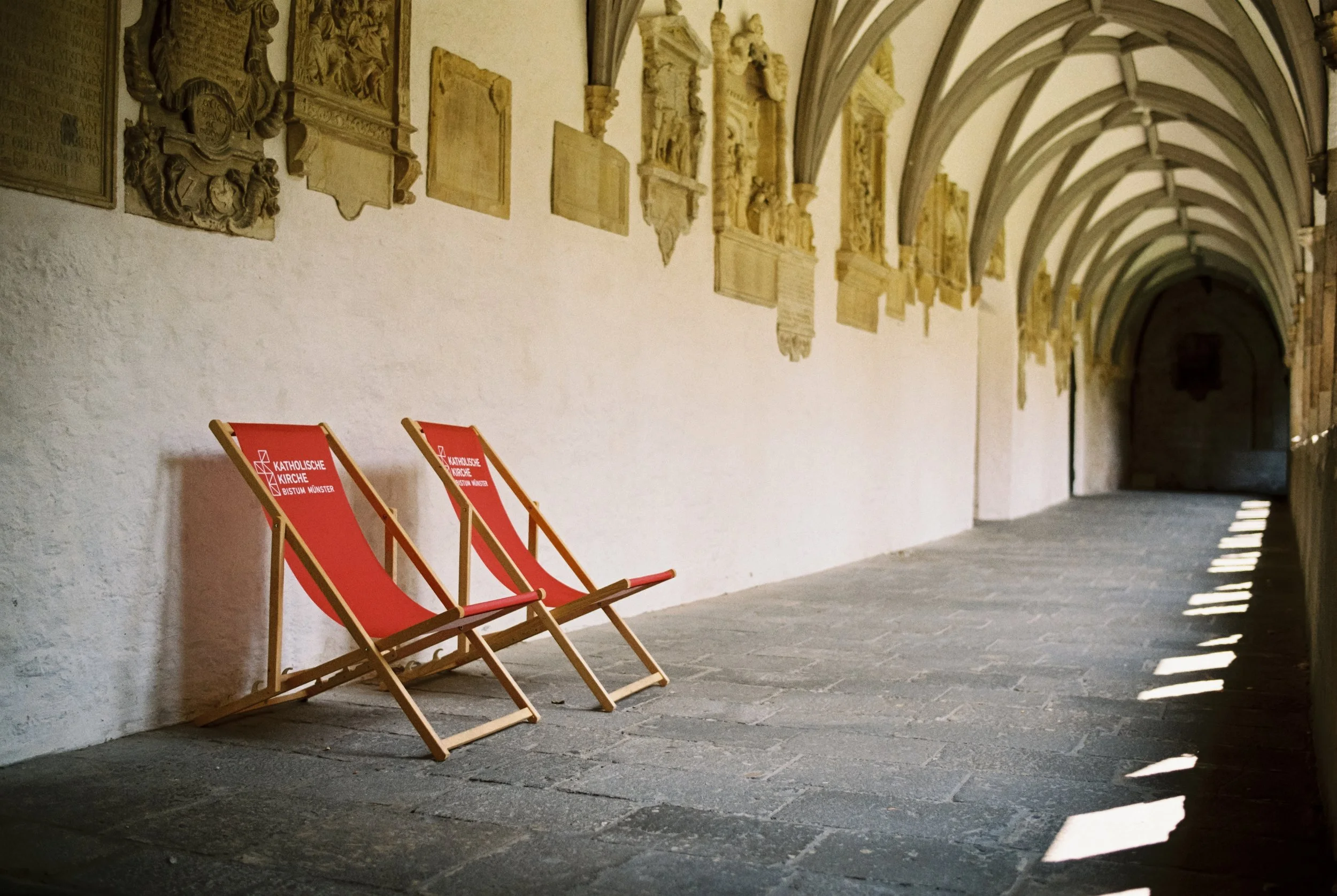 Zwei rote Liegestühle mit dem Logo der Katholischen Kirche und des Bistums Münster, in einer mittelalterlichen Arkadenhalle mit Gemälden an der Wand und Sonnenlicht, das auf den Boden scheint.