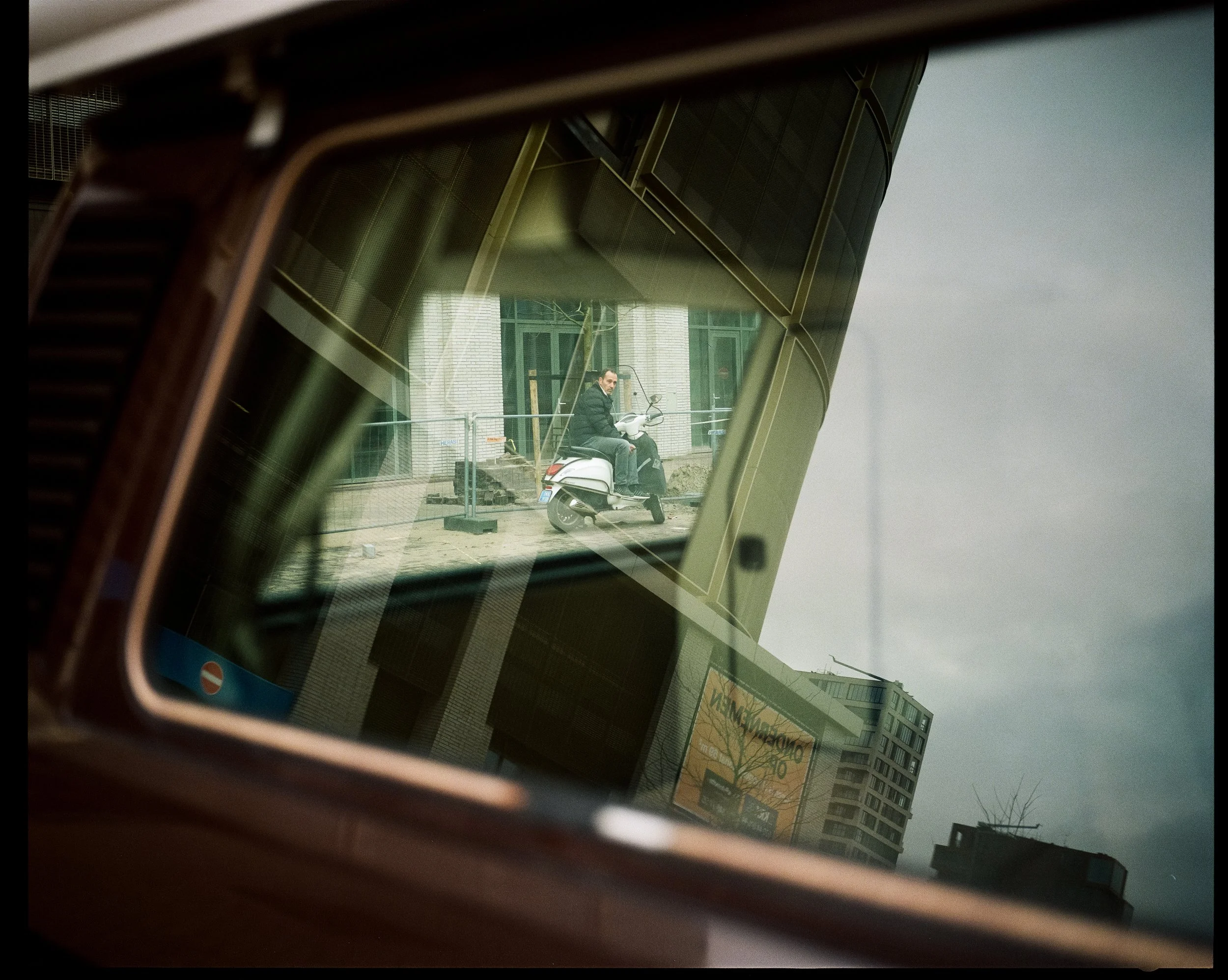 Reflexion eines Mannes auf einem weißen Motorroller, die in einem Fenster eines Gebäudes reflektiert wird. Im Hintergrund sind urbane Gebäude und ein grauer Himmel sichtbar.