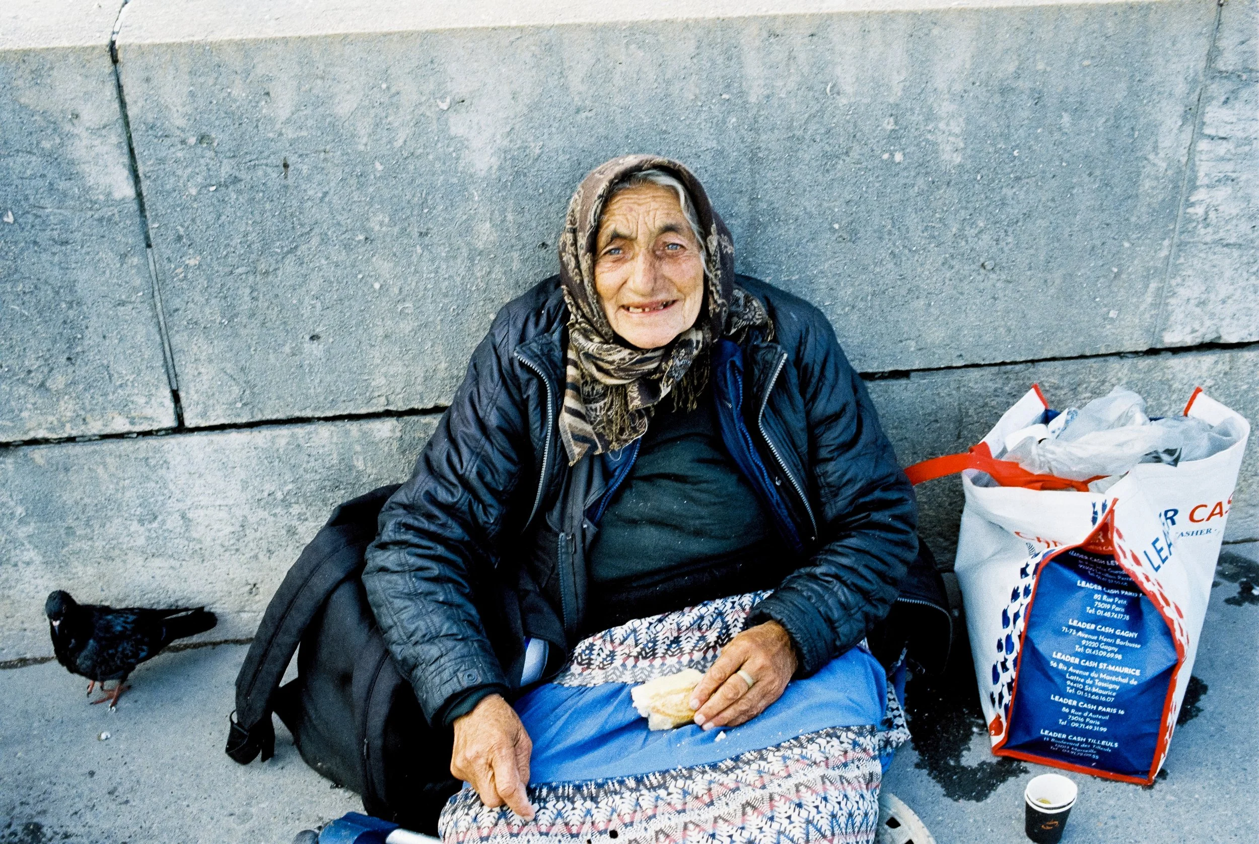 Obdachlose Frau sitzt auf der Straße vor einer Steinmauer, hat eine Einkaufstüte neben sich und isst ein Brötchen. Es ist eine Krähe in der Nähe.