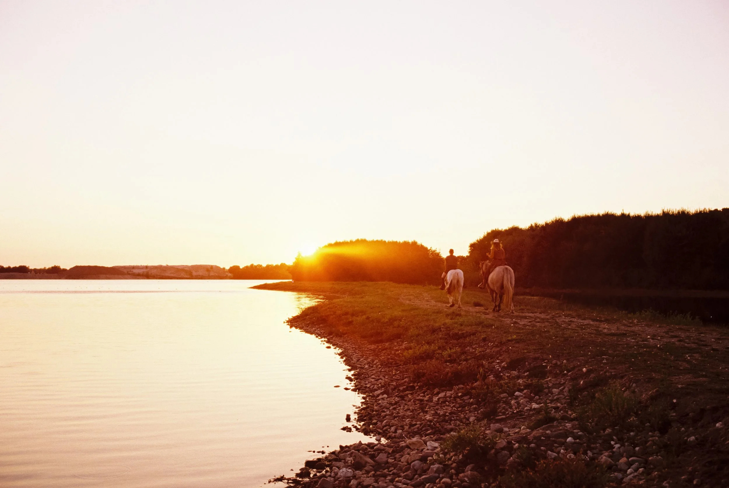 Zwei Reiter auf Pferden reiten entlang eines Flusses bei Sonnenuntergang, umgeben von Büschen und einer ruhigen Wasserfläche.