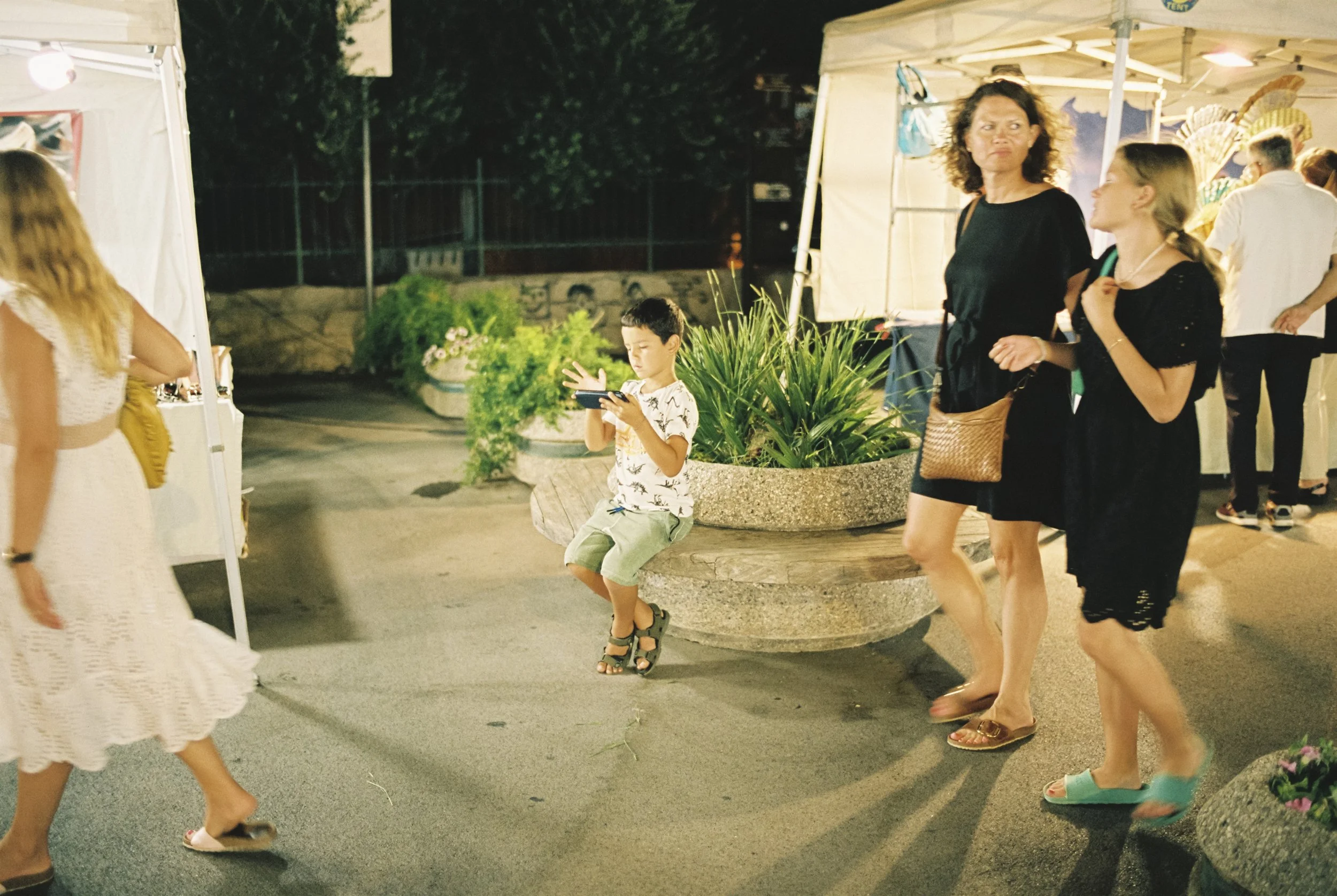 Eine Gruppe von Menschen schlendert nachts an einem Marktstand mit geöffneter Markise vorbei, im Hintergrund sind große Pflanzkübel mit grünen Pflanzen und eine dunkle Felsenmauer sichtbar.