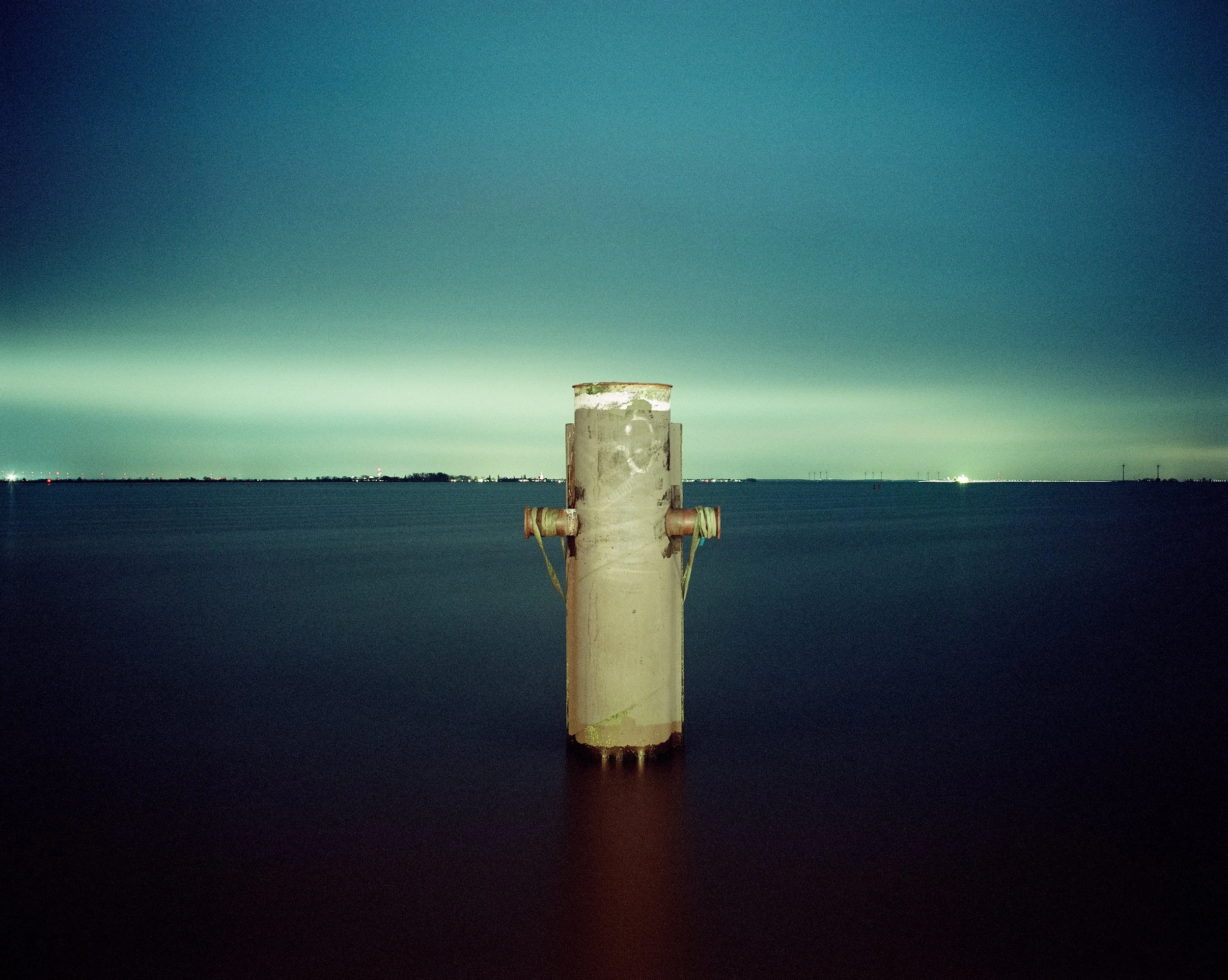 Ein rostiger Pfosten im Wasser mit einer Stadt- oder Industrieansicht im Hintergrund bei Nacht.