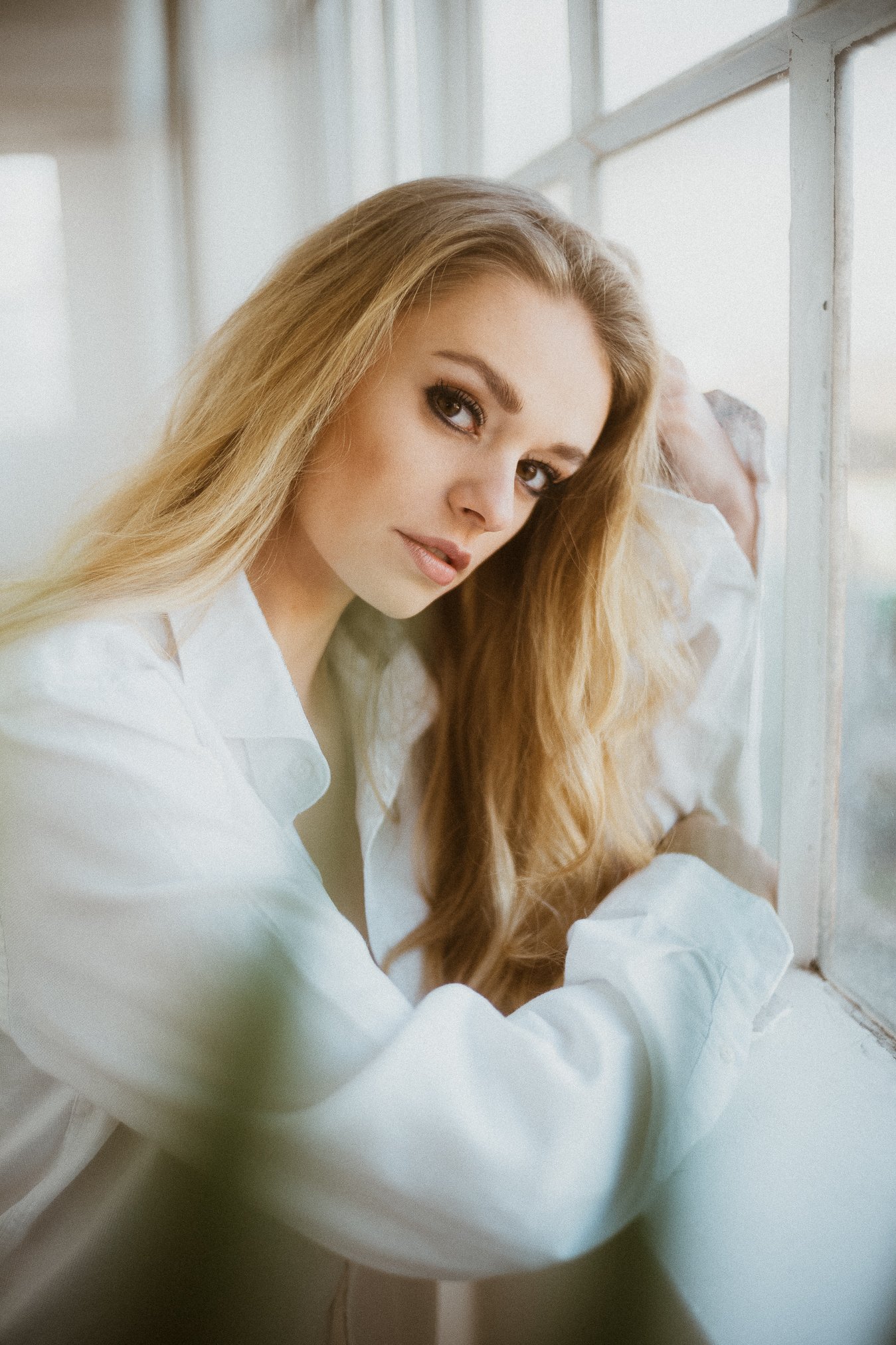 Eine junge Frau mit langen, lockigen blondem Haar und hellbraunen Augen, die neben einem Fenster sitzt und in die Kamera schaut.