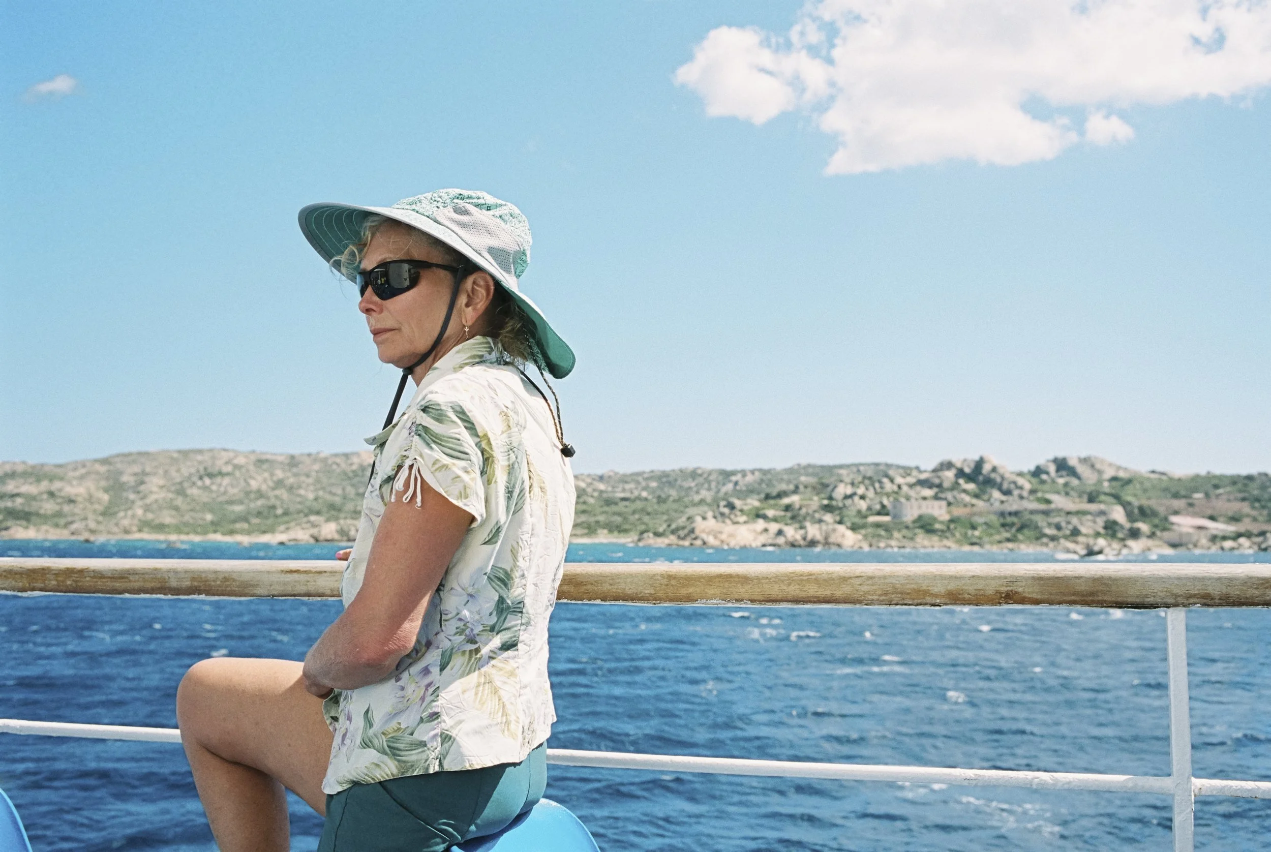 Eine ältere Frau trägt einen Hut, Sonnenbrille und Sommerkleidung, sitzt auf einem Boot mit Blick auf das Meer und eine Insel im Hintergrund.