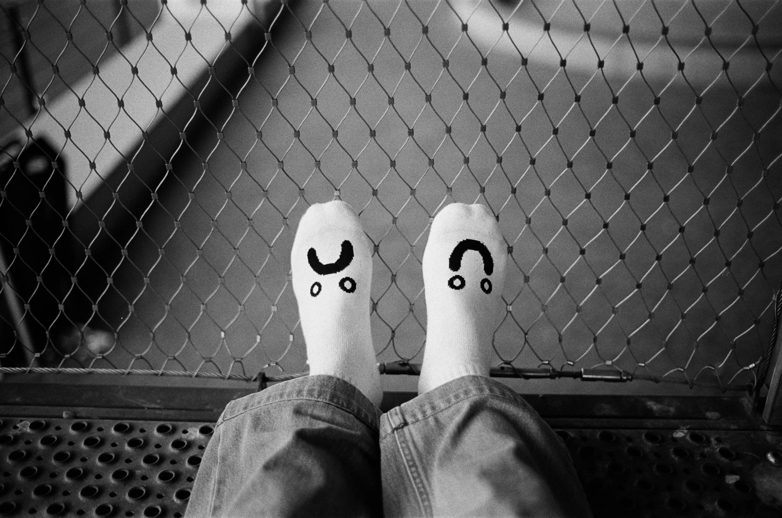 Blick nach unten auf eine Person in Jeans mit weißen Socken, die auf einer Metallplattform steht, die Füße vor einem Drahtzaun, mit lachenden Gesichtern auf den Socken.