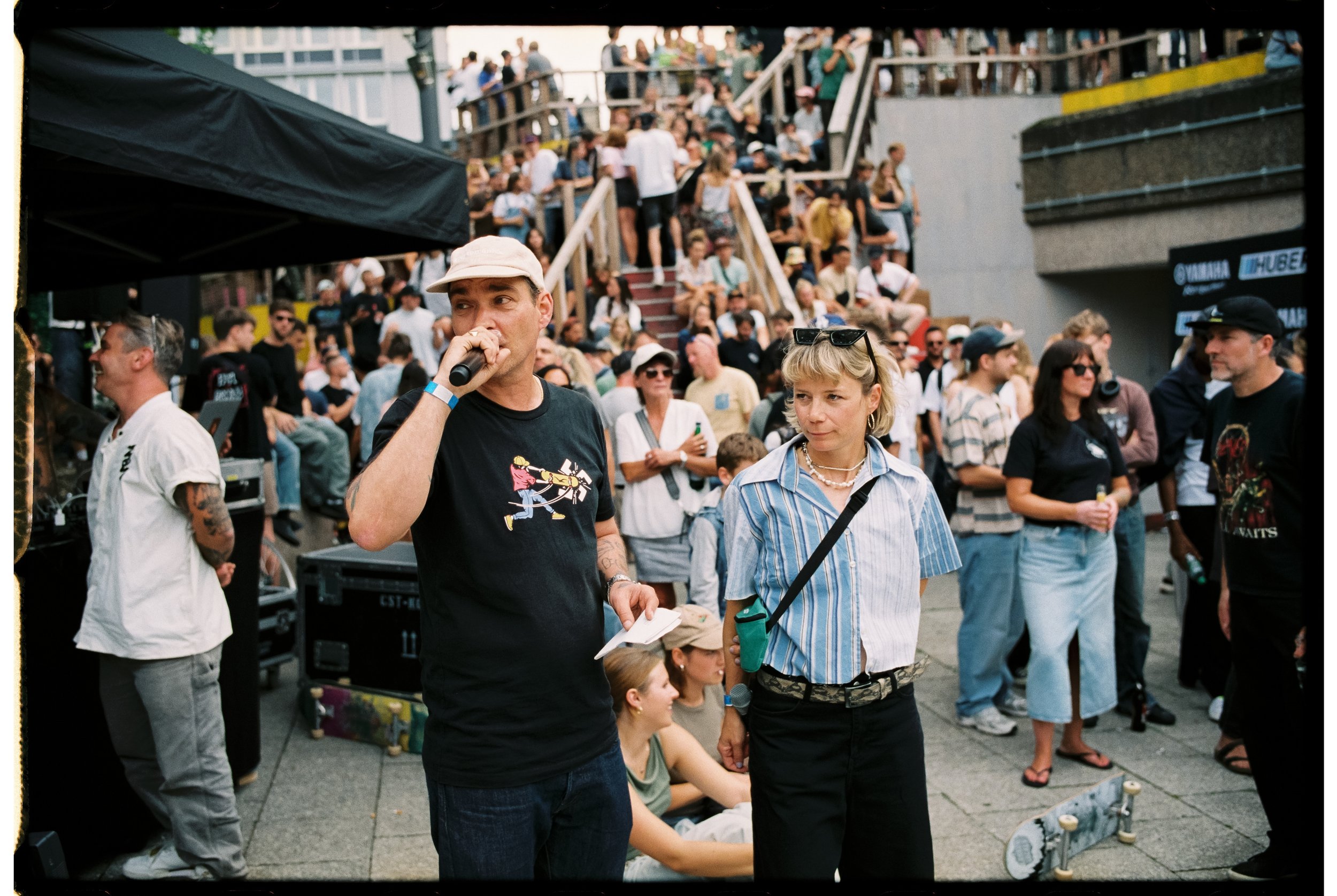 Menschen versammeln sich bei einem Musik- oder Kulturfestival im Freien, einige stehen, andere sitzen auf Stufen, im Hintergrund ist eine Bühne oder ein Bereich mit Snack- und Getränkeständen sichtbar. Im Vordergrund spricht ein Mann mit Mikrofon, ne