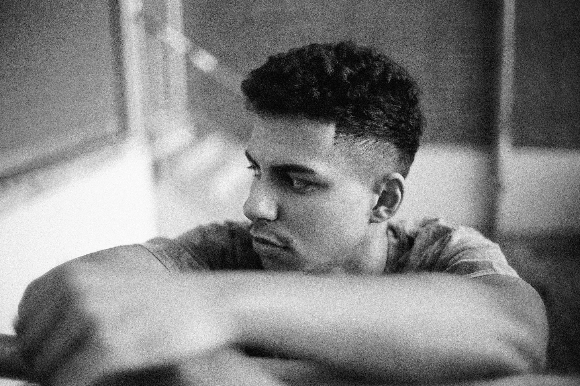 Schwarz-weiße Aufnahme eines jungen Mannes mit kurzen, lockigen Haaren, der nachdenklich nach links schaut mit seinem Arm auf einem Tisch oder einer Oberfläche.