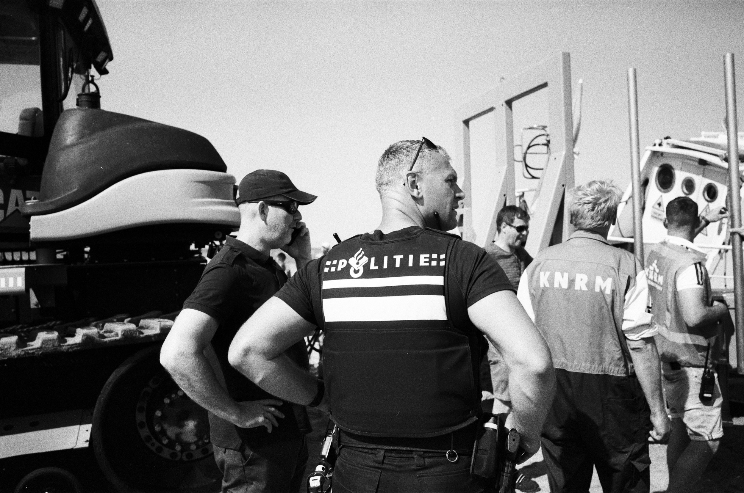 Polizeibeamte stehen auf einer Straße, einige sprechen, andere schauen auf die Ausrüstung. Im Hintergrund sind ein Fahrzeug und eine größere Bau- oder Befestigungsstruktur. Es herrscht Tagessonne, Bild in Schwarzweiß.