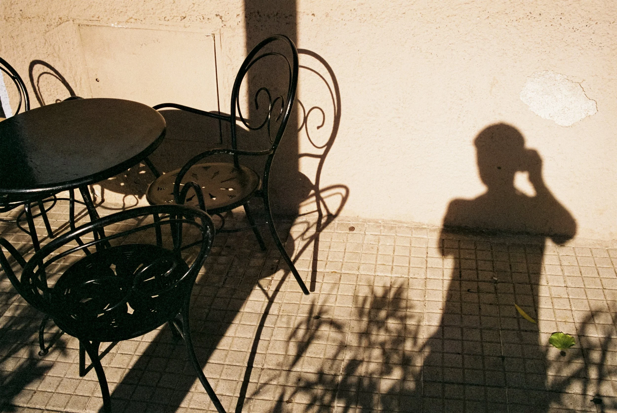 Schatten einer Person, die ein Smartphone hält, auf einer beige Wand, neben schwarzen Metallmöbeln auf einem Fliesenboden bei Sonnenlicht.