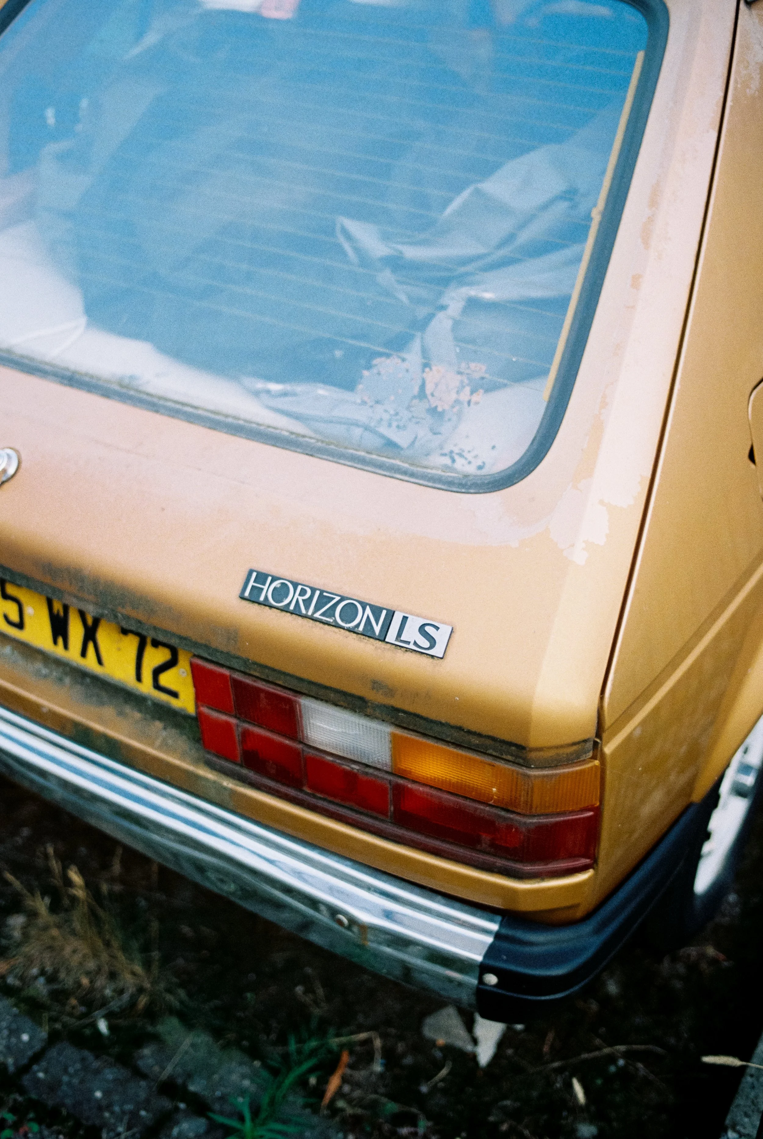 Das Bild zeigt die Rückseite eines gelb-orangefarbenen Autos mit einem Fenster, das mit Mülltüten bedeckt ist. Es trägt ein Abzeichen mit der Aufschrift 'HORIZON LS'.