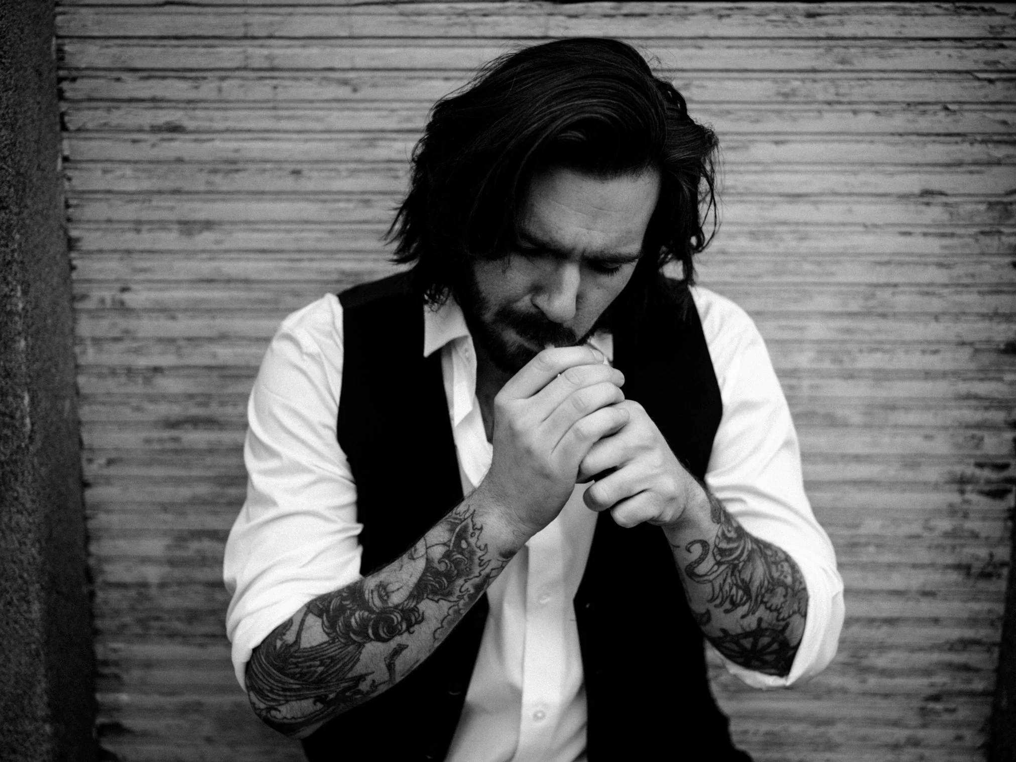 Schwarz-weiß-Foto eines Mannes mit Tätowierungen auf den Armen, mit langen Haaren, der nachdenklich seine Hände vor den Mund hält, vor einer Holzwand.