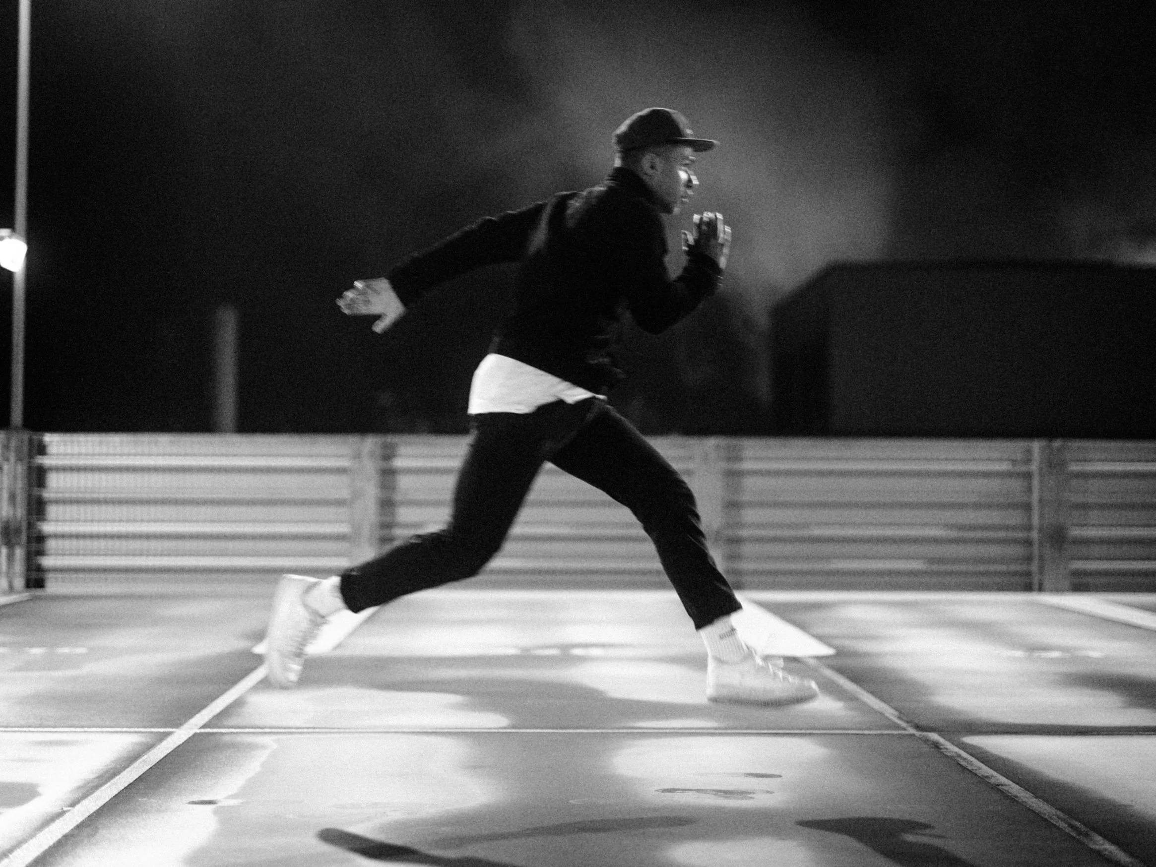 Ein Mann läuft nachts auf einem Parkplatz, trägt einen Hut, eine dunkle Jacke, weiße Schuhe und hat seine Hände vor den Mund gehalten, als ob er schriebe oder rufe.