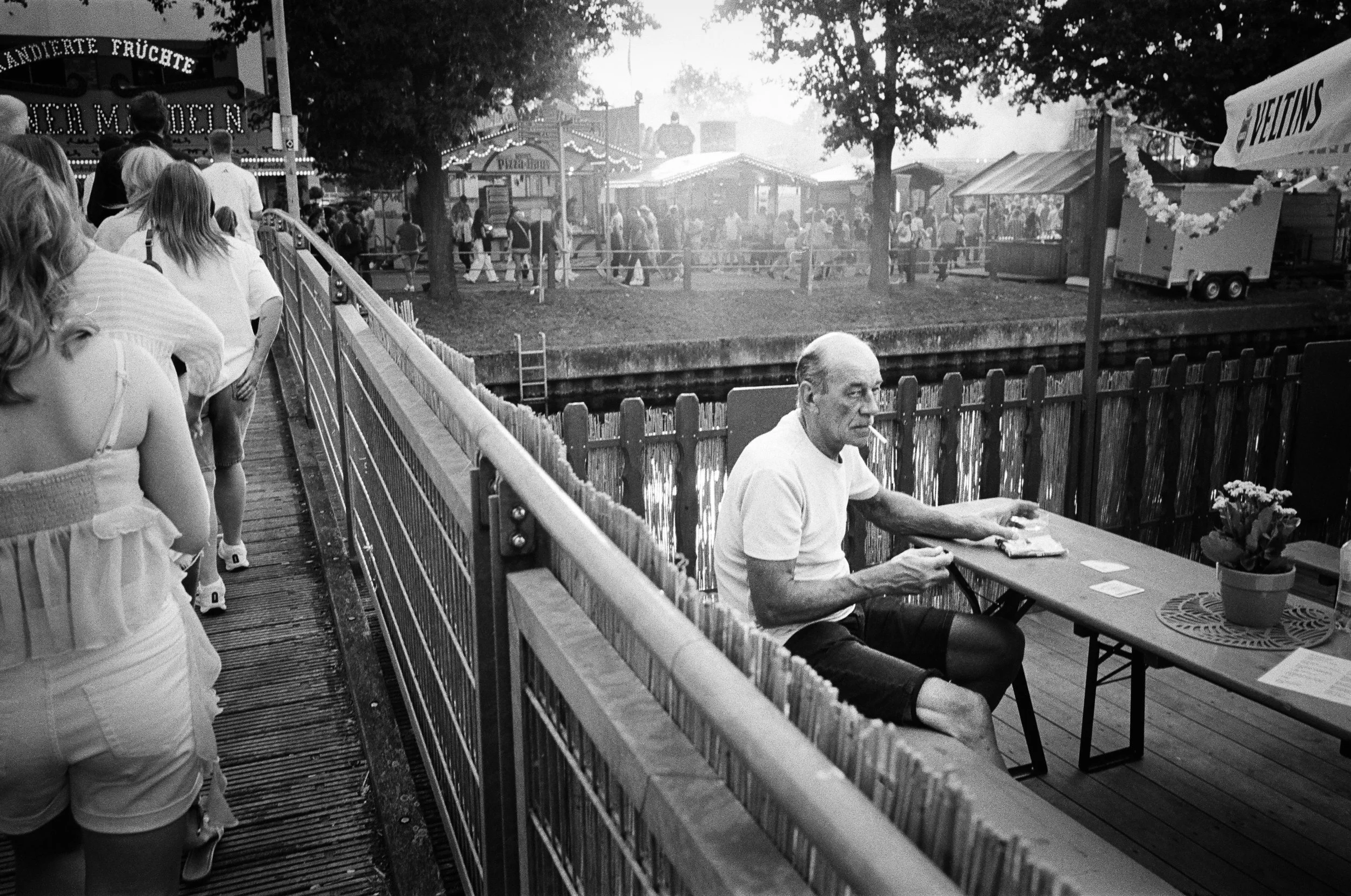 Ein älterer Mann sitzt an einem Tisch neben einem Wasserkanal, mit Zigaretten im Mund, während Menschen in einer Menschenmenge auf einem Festplatz oder Jahrmarkt unterwegs sind.