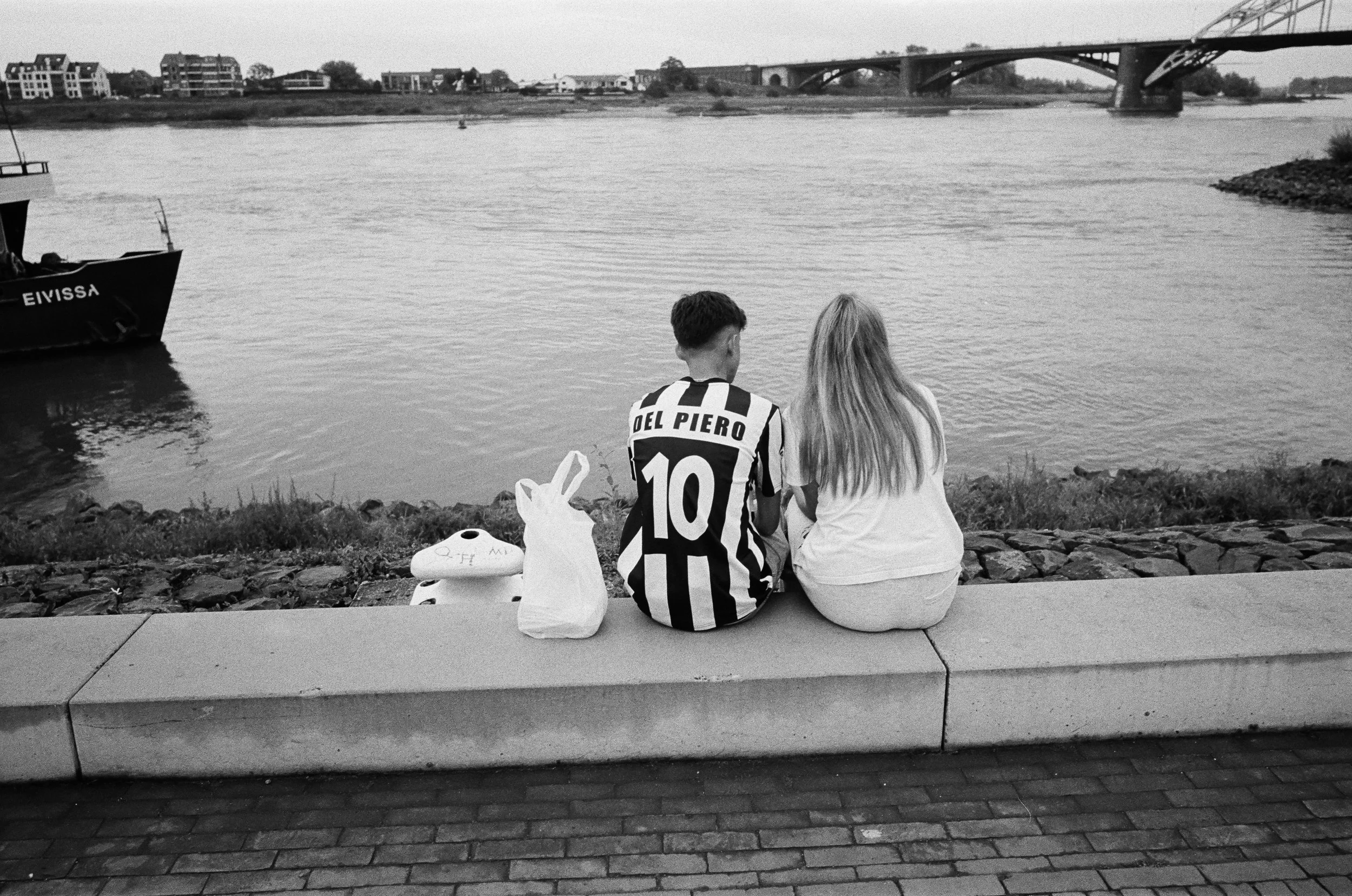 Ein junger Mann und eine junge Frau sitzen am Ufer eines Flusses, mit ihren Rücken zum Betrachter. Der Mann trägt ein schwarz-weiß gestreiftes Fußballtrikot mit der Nummer 10 und dem Namen 'DEL PIERO'. Neben ihnen liegt eine Tasche und eine weiße Tüt