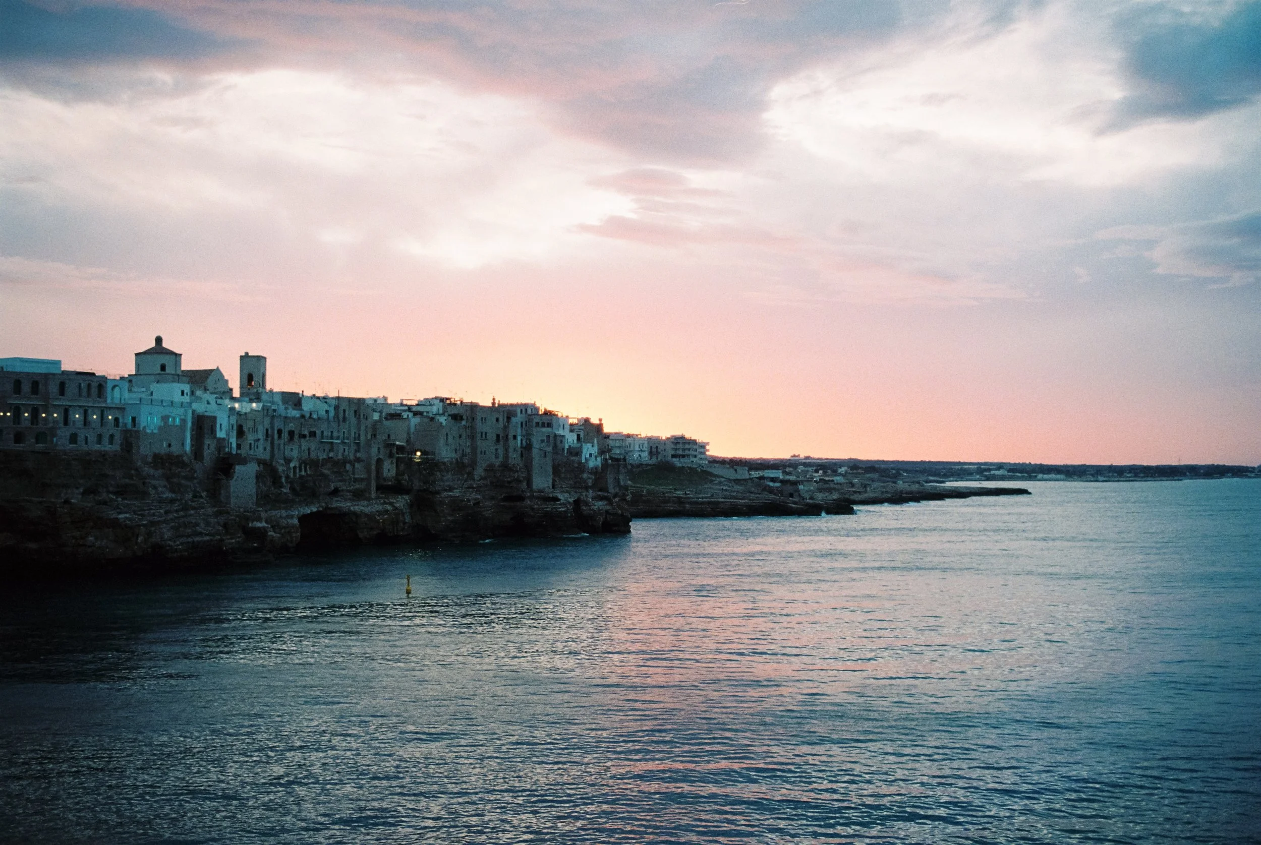 Blick auf eine Küstenstadt bei Sonnenuntergang, mit weißen Gebäuden auf Klippen und ruhigem Wasser im Vordergrund.