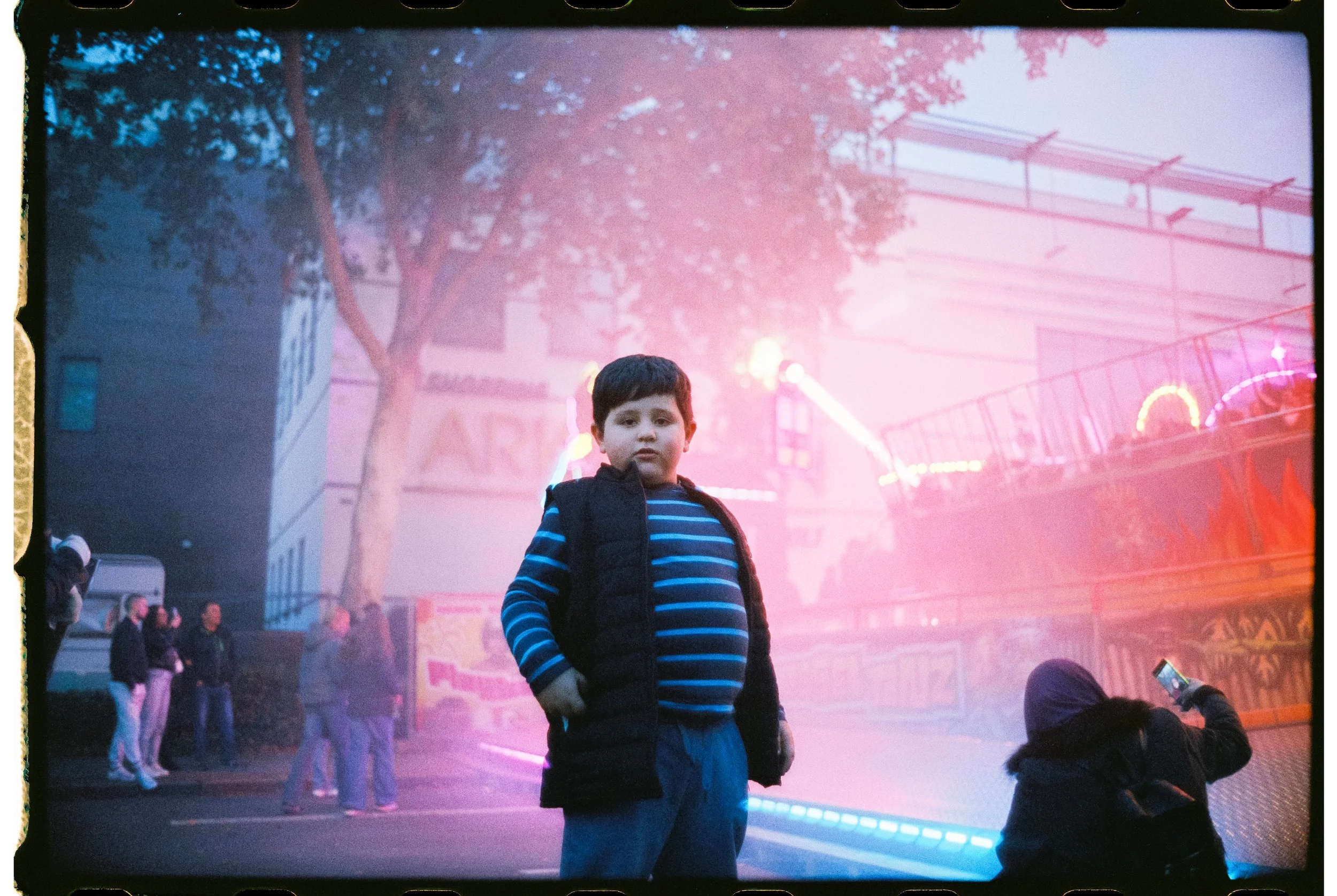 Ein Junge steht vor einem Jahrmarkt mit leuchtenden Fahrgeschäften, während im Hintergrund andere Menschen stehen und ein Mädchen ein Foto macht.