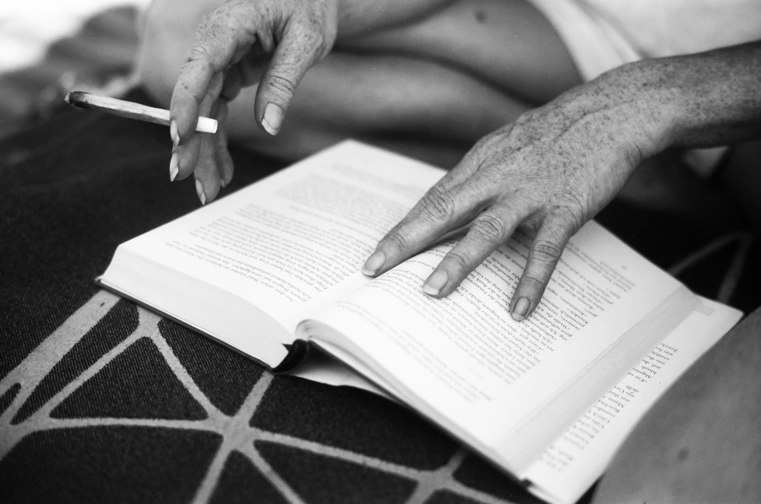 Nahaufnahme von älteren Händen, die in einem Buch lesen und einen Stift halten. Das Buch liegt auf einem geometrisch gemusterten Stoff.