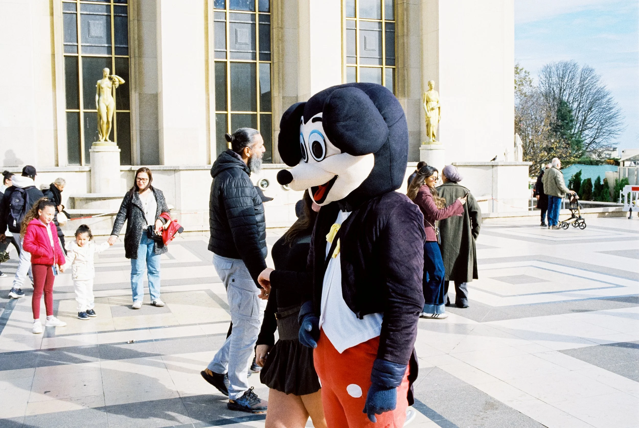 Person in einem Minni Maus Kostüm posiert vor einem historischen Gebäude mit Statuen, während andere Menschen im Hintergrund im Sonnenschein spazieren gehen.