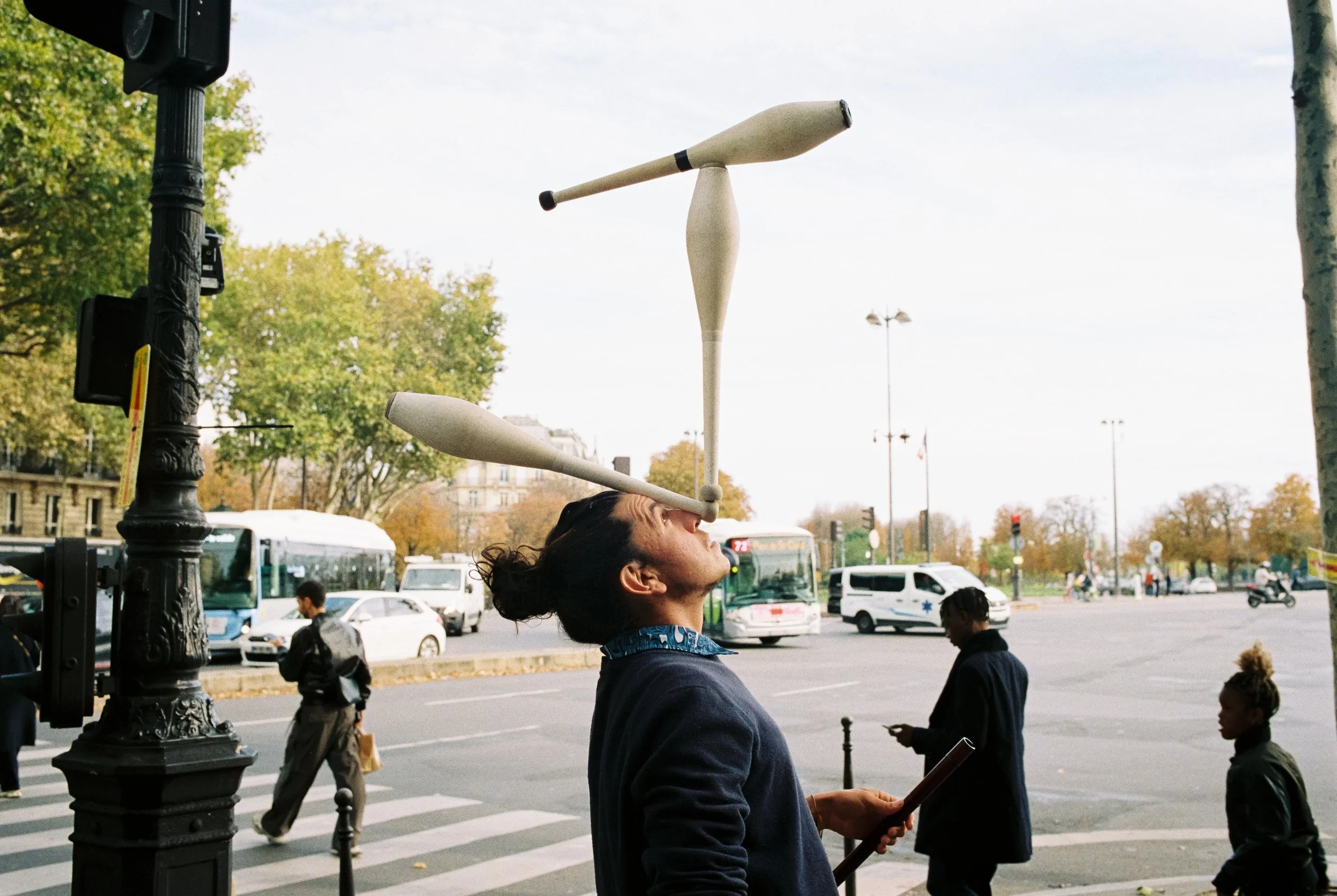 Ein Mann balanciert zwei Holzstäbe auf seinem Gesicht, eine auf der Stirn und eine auf der Nase, während er auf einer Straße in einer Stadt steht.